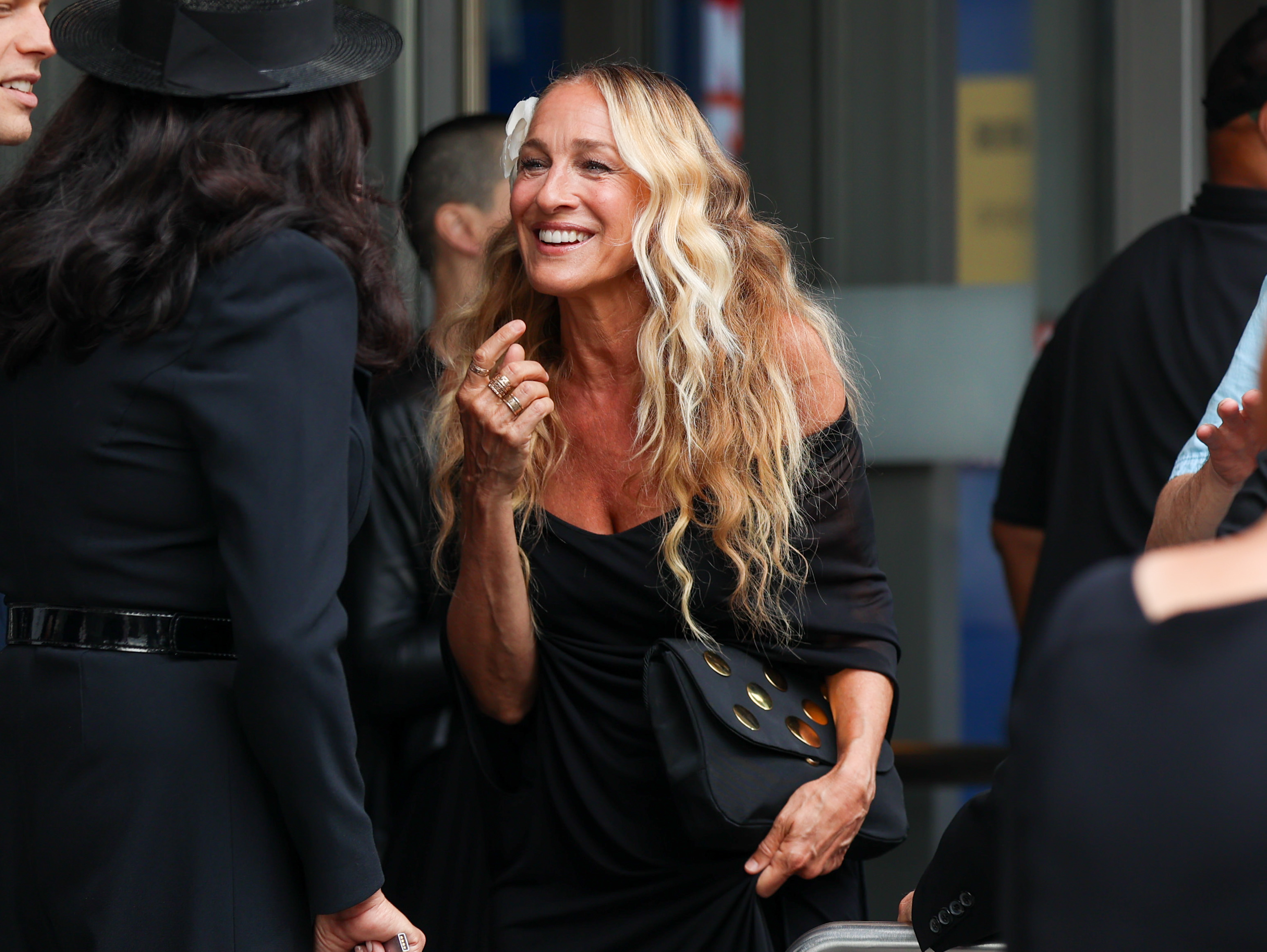 Sarah Jessica Parker is smiling and talking to a person in a hat at an event, wearing a chic off-the-shoulder black dress and holding a black bag with gold accents