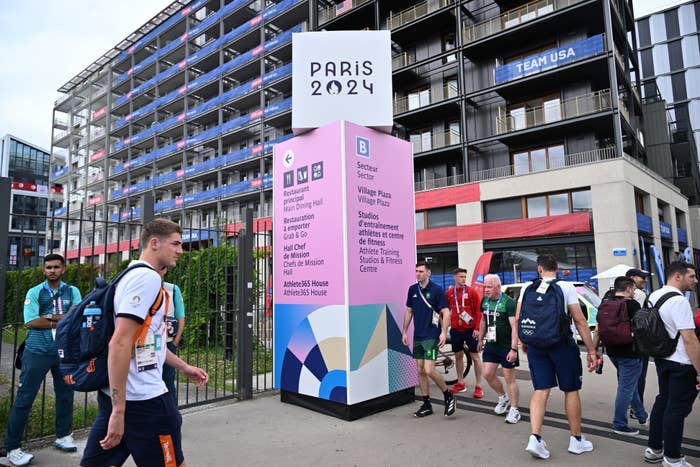 Athletes and officials walk in front of a sign for the Paris 2024 Olympics at the Athlete's Village Plaza. The sign lists various facilities and locations