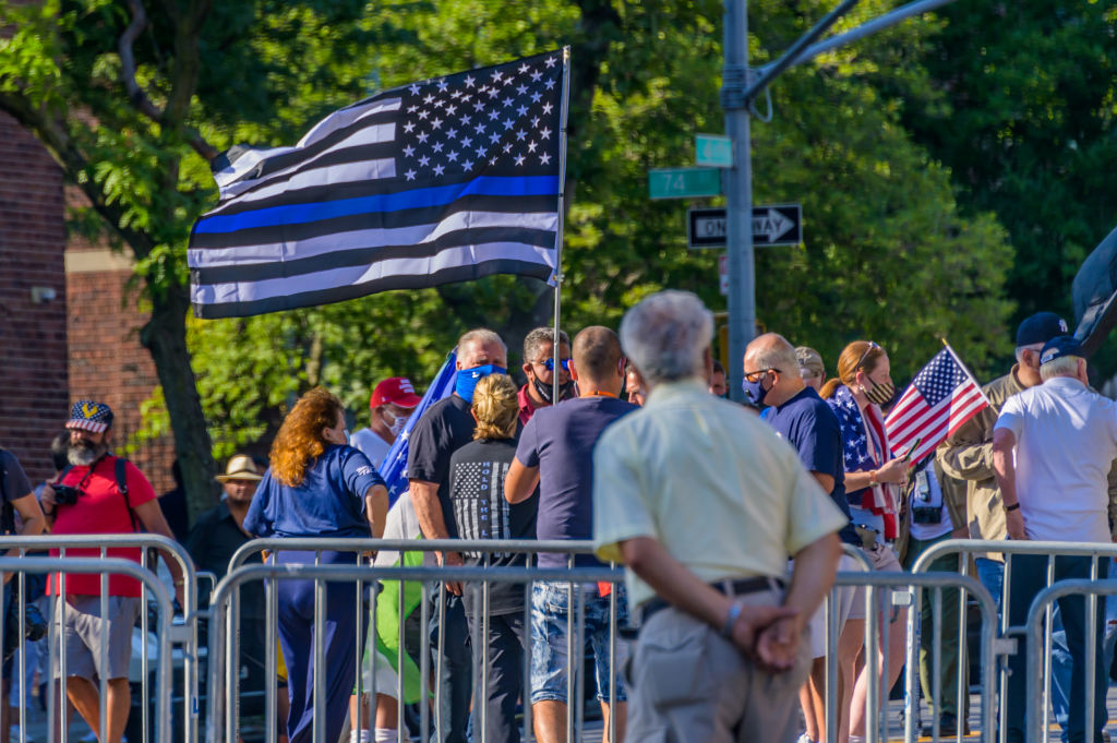 People gather at a rally holding American flags and a pro-police thin blue line flag, standing behind a barricade, discussing an event