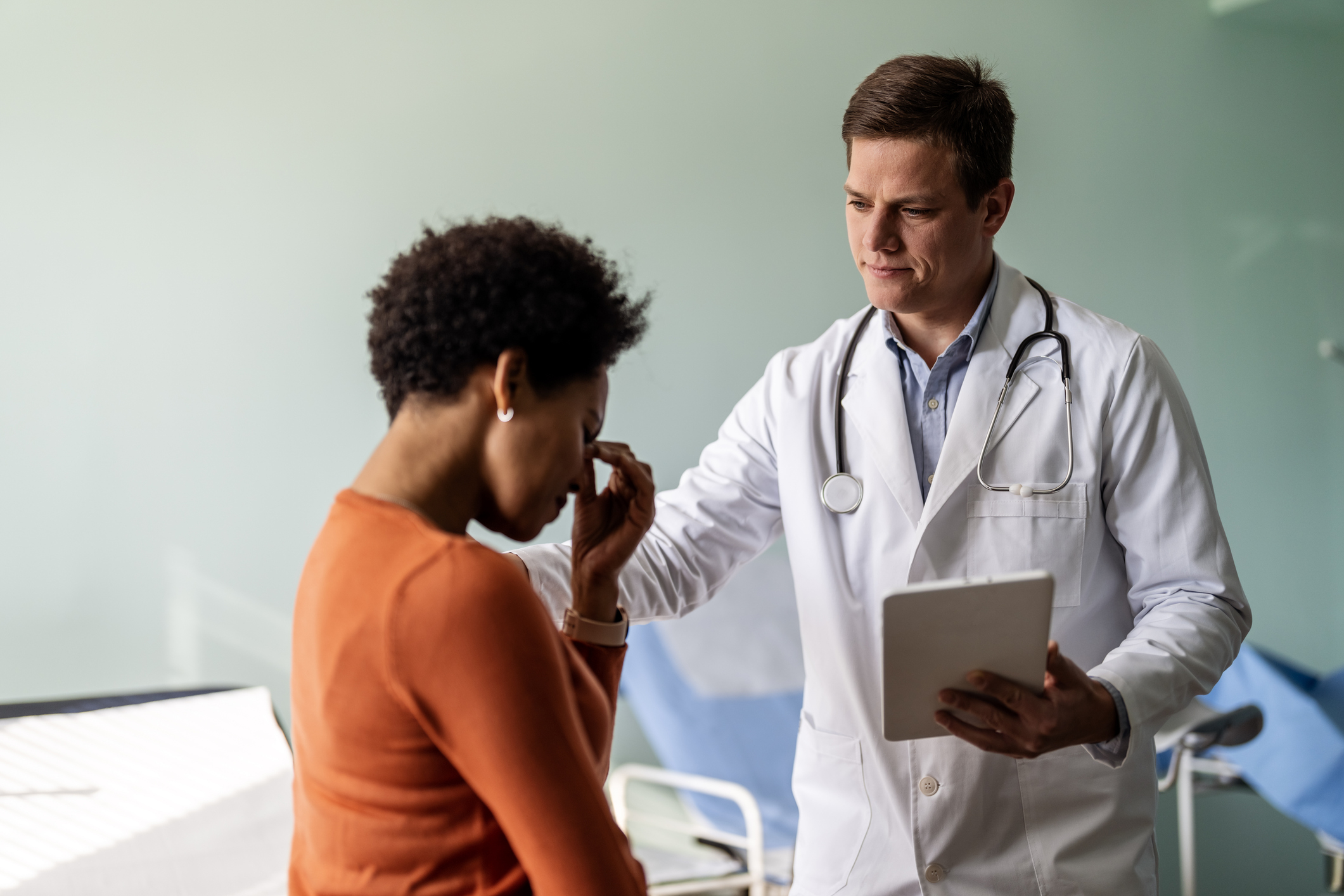 A doctor, wearing a white coat and holding a tablet, comforts a woman who appears distressed in a medical office setting