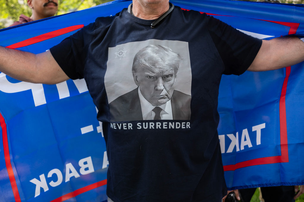 A person is wearing a shirt with Donald Trump's face and the words &quot;Never Surrender&quot; showing a stern expression. They are holding a blue flag in the background