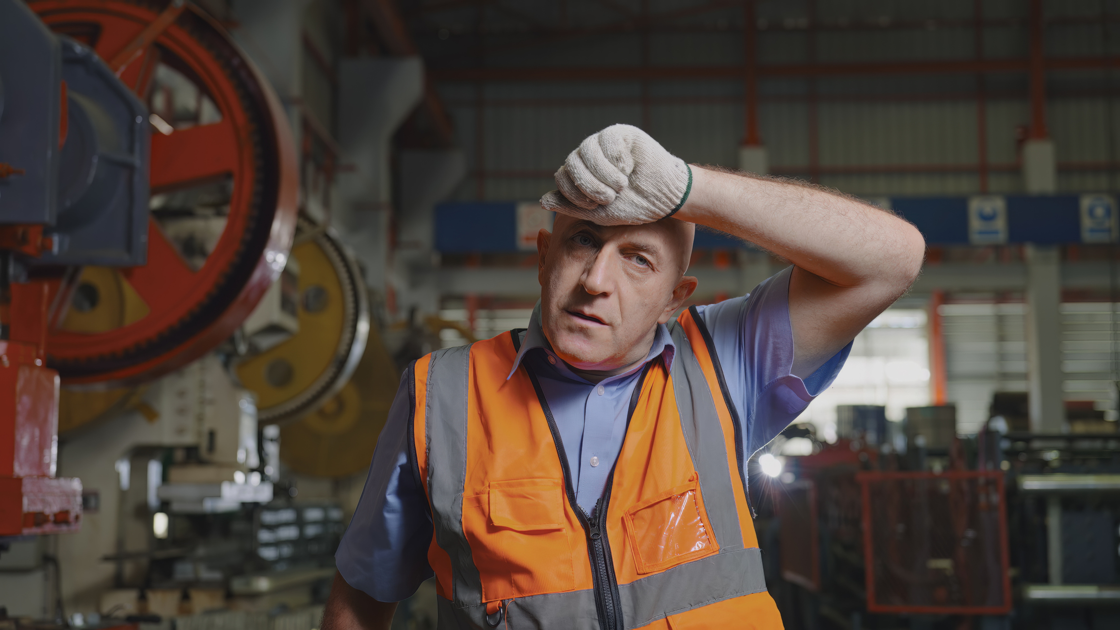 A factory worker in a safety vest wipes sweat from his forehead inside a manufacturing plant, surrounded by industrial machinery