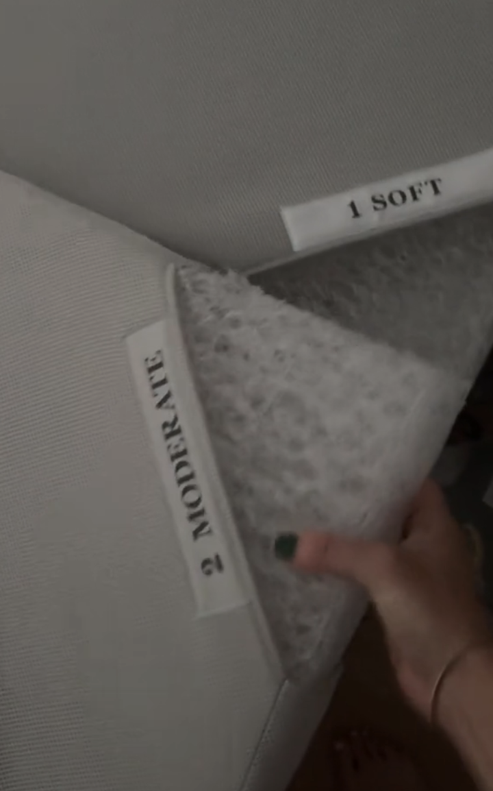 A hand lifting a mattress corner to show two labels: "1 SOFT" and "2 MODERATE," indicating different levels of firmness