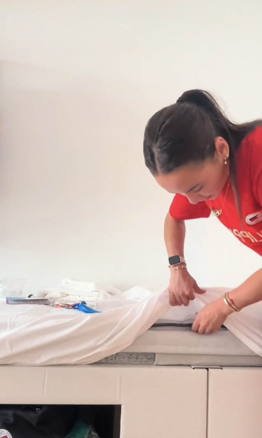 A woman in a red shirt is putting a white cover on a mattress. Various items are on the bed
