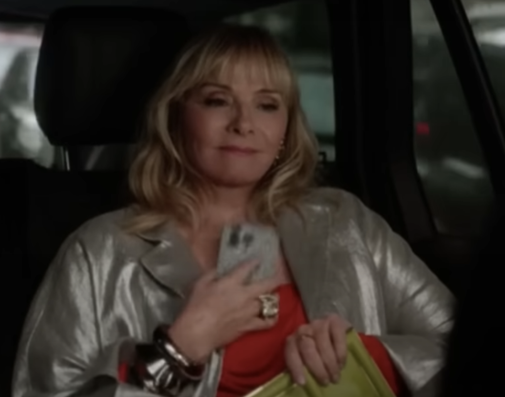 Kim Cattrall sits in a car, holding a phone and a yellow clutch, wearing a metallic jacket over a red top