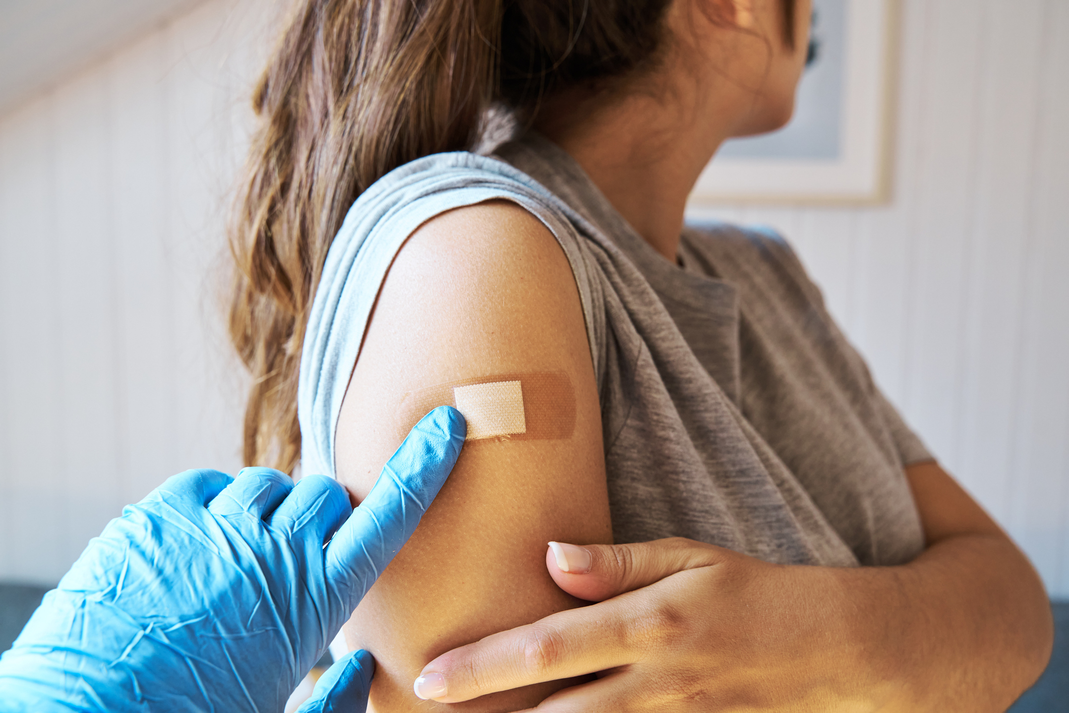 A healthcare worker wearing blue gloves applies a bandage to a woman's upper arm after a vaccination. The woman is looking away