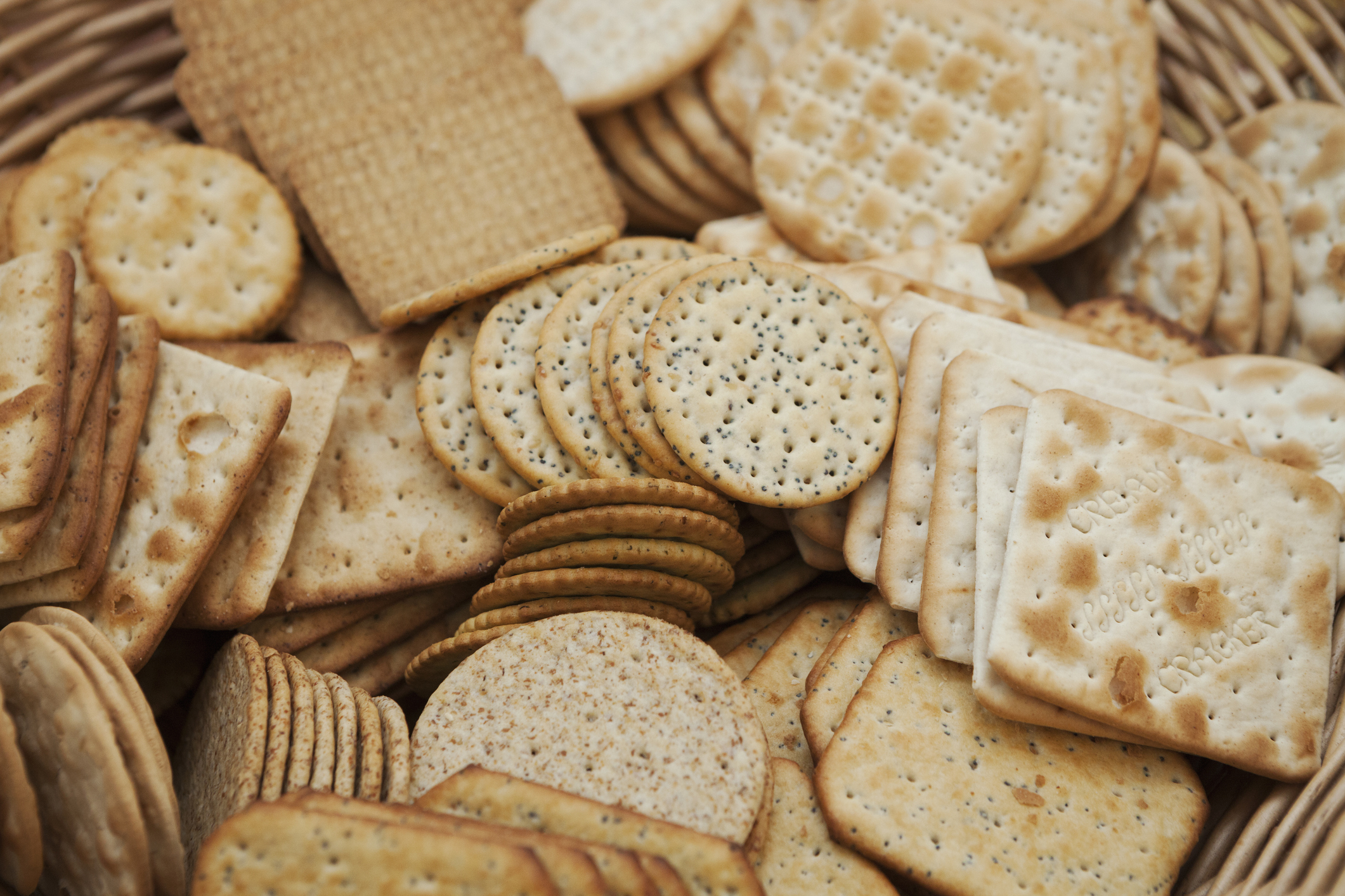 A variety of crackers arranged in a woven basket, including round, square, and rectangular shapes with different textures and toppings
