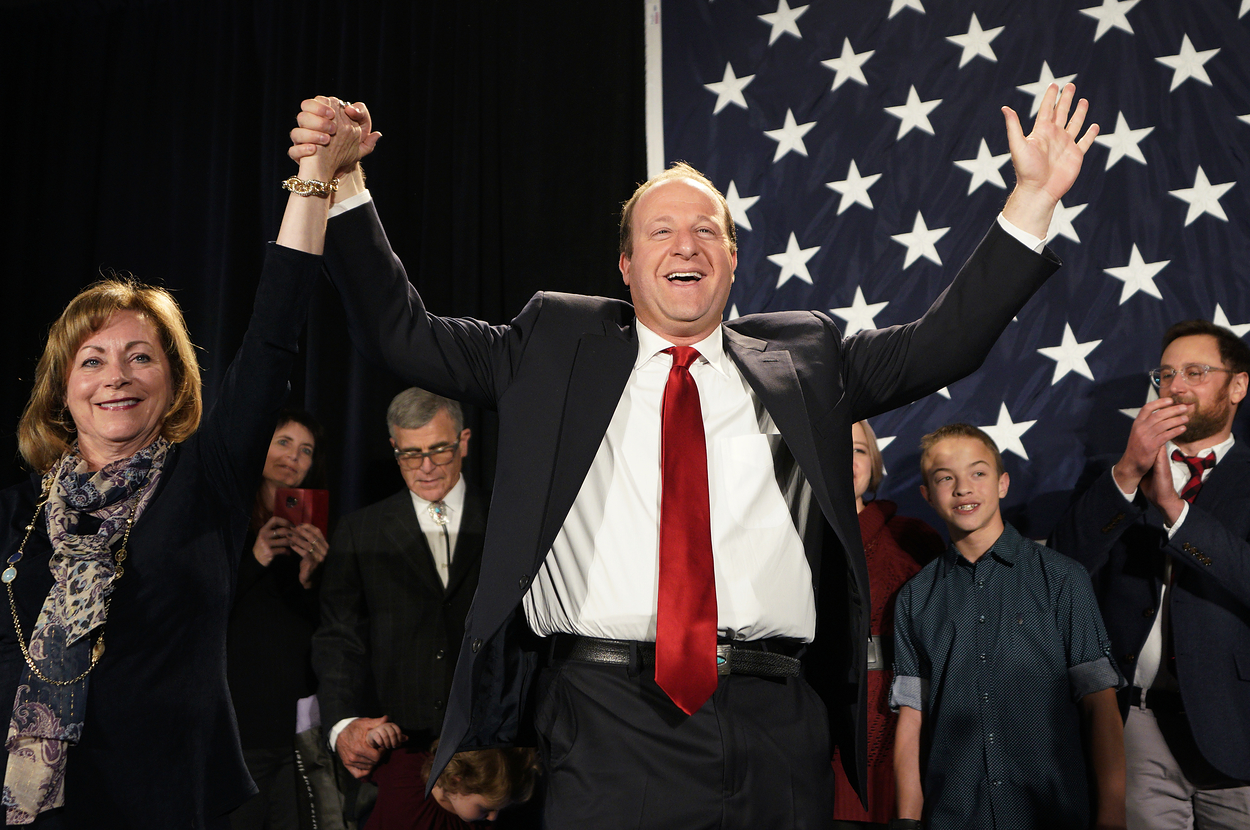 Jared Polis and Marlon Reis celebrate with supporters in front of a starred flag backdrop. Jared holds up hands with a woman beside him