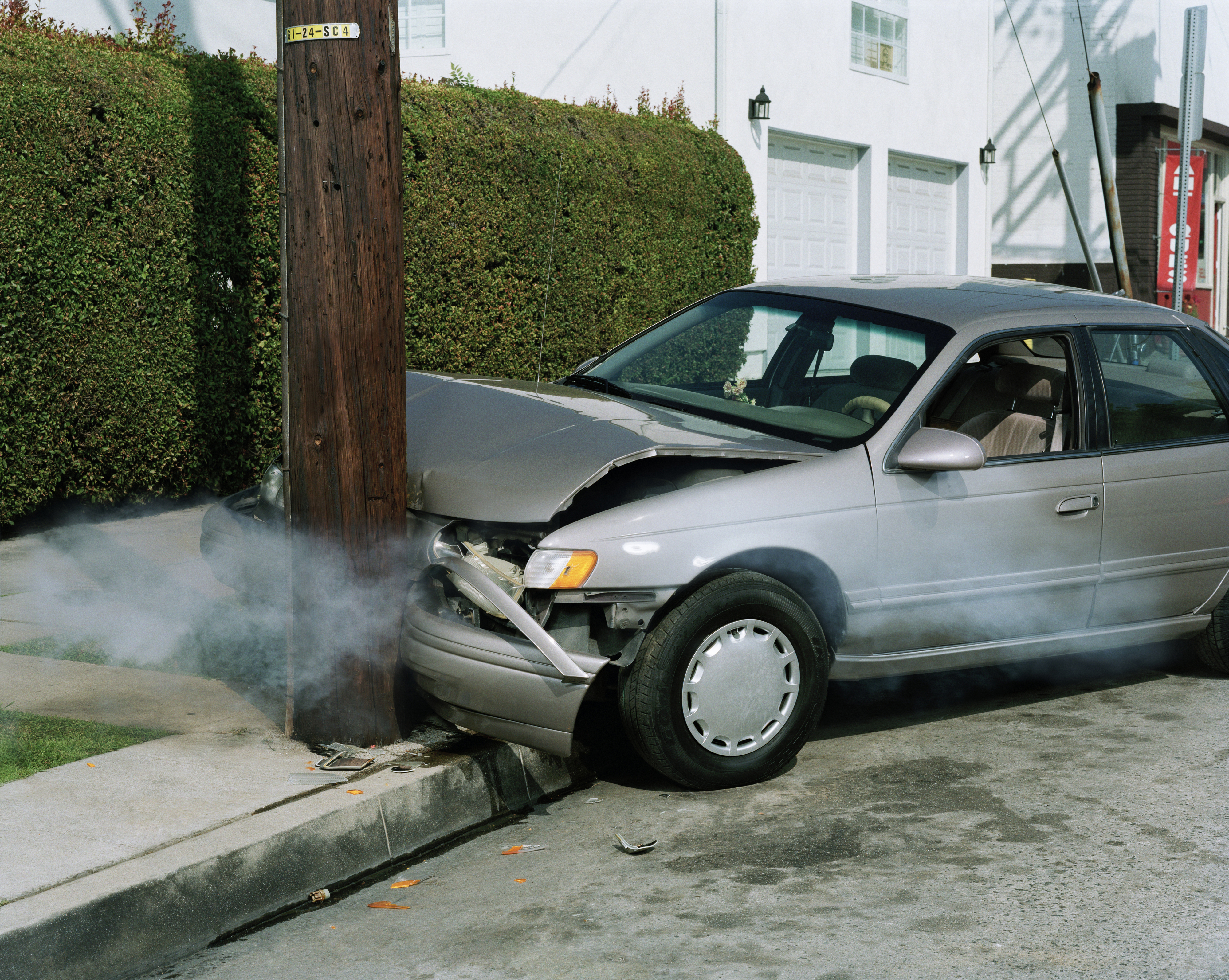 A car has crashed into a utility pole on a suburban street, with smoke emerging from the vehicle’s front. There are no people visible in the image