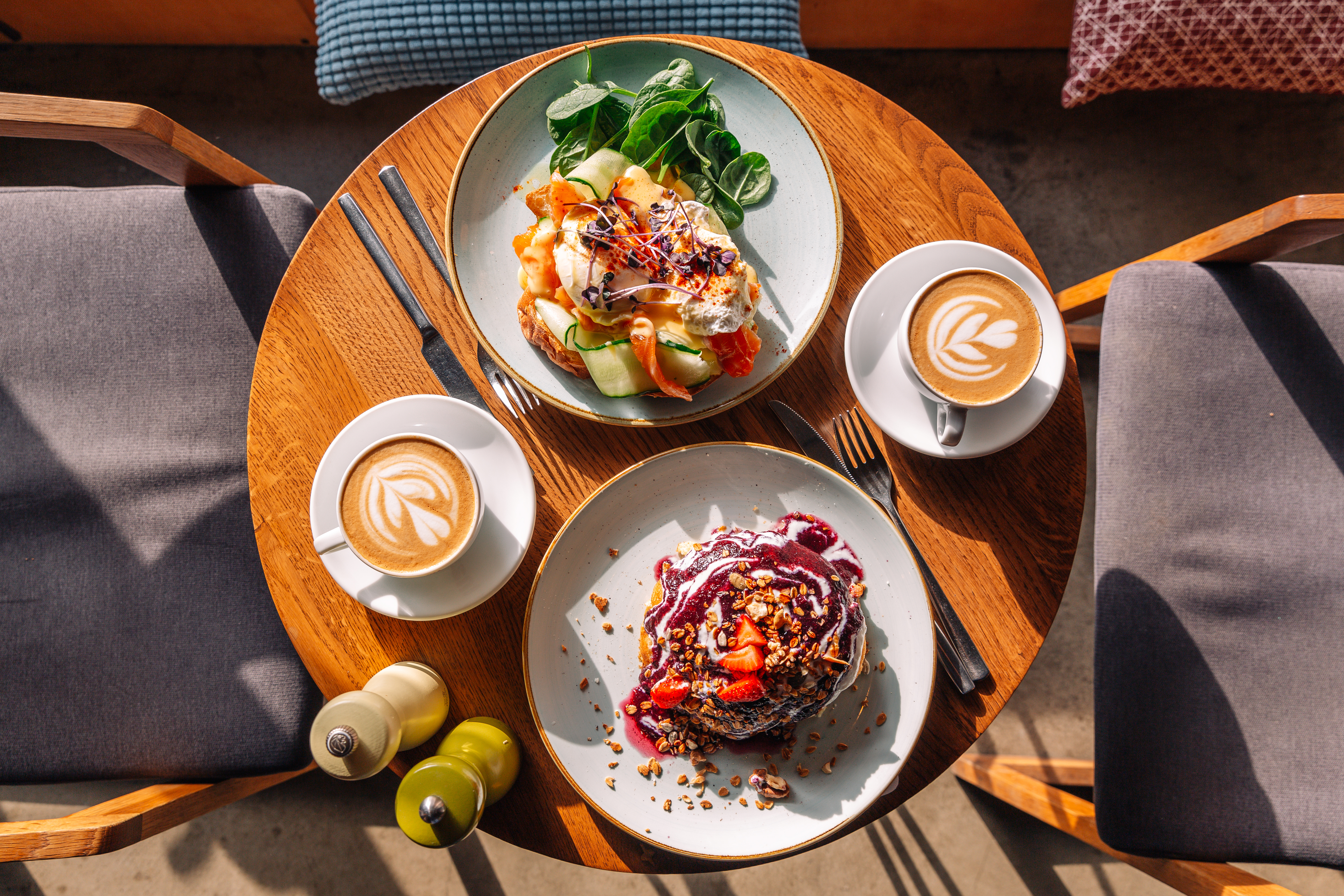 A round wooden table with two plates of gourmet food, two lattes with latte art, and salt and pepper shakers. The table is set for two