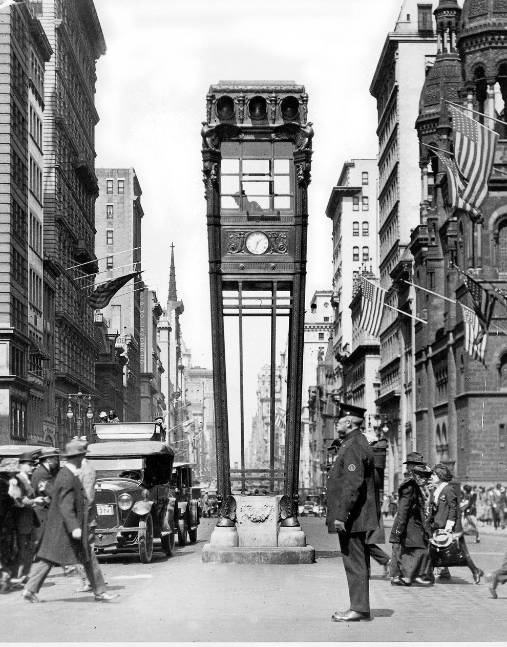 Vintage photograph of New York City's 5th Avenue with a tall clock structure in the center of the street. The street is bustling with people and old cars