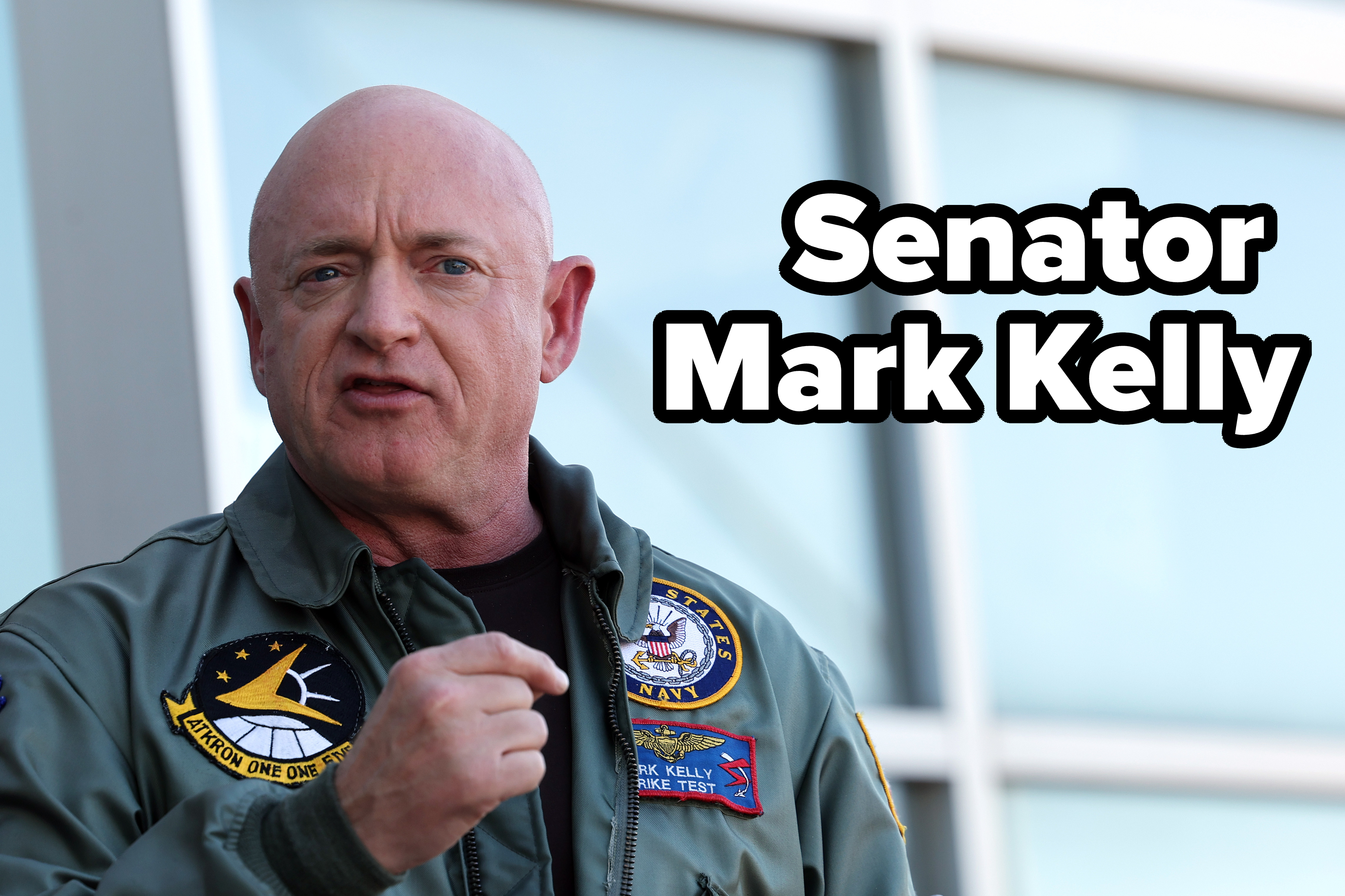 Mark Kelly speaks while wearing a flight suit with various patches, including a NASA patch and a U.S. Navy insignia, in front of a modern building