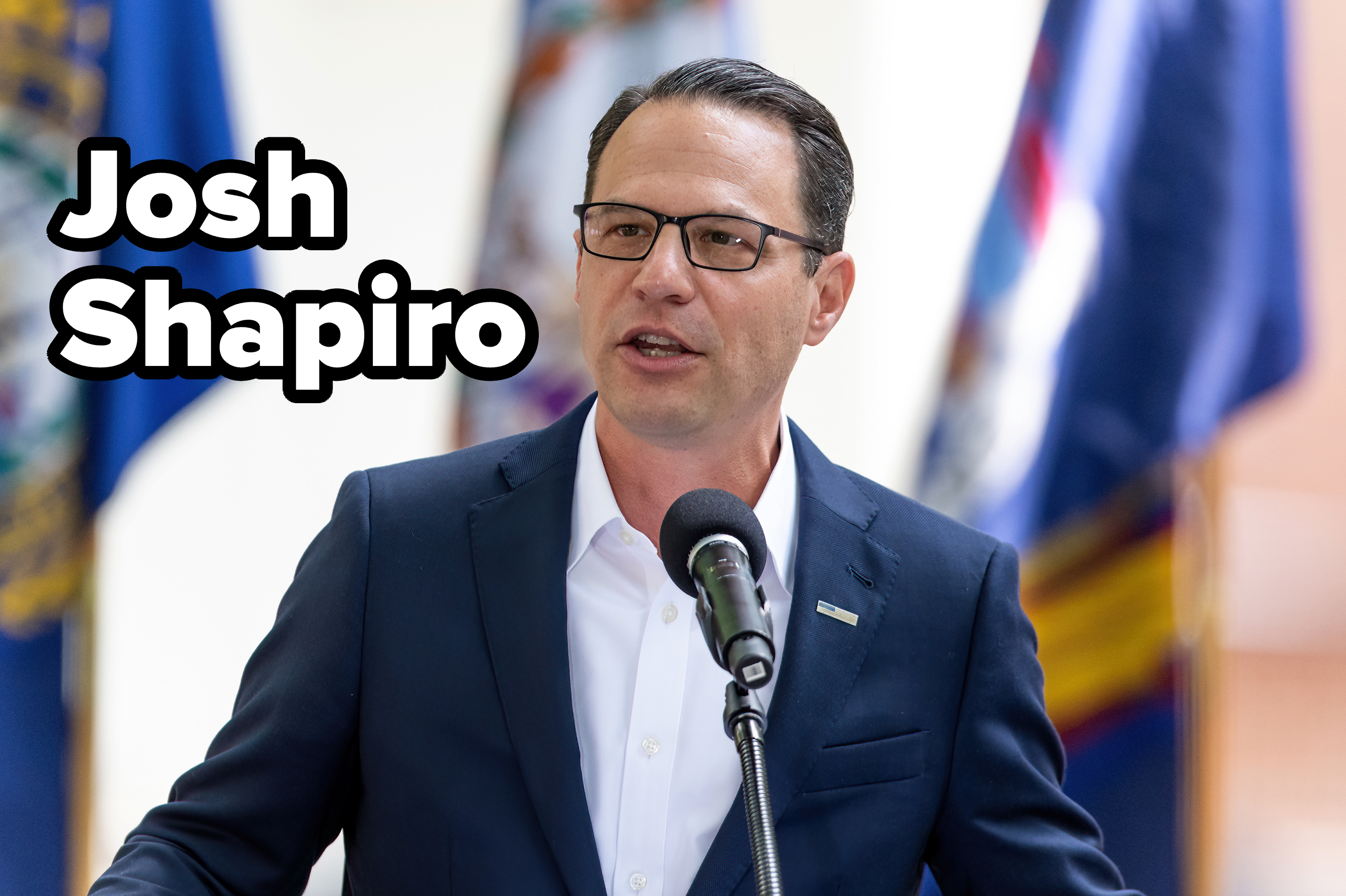 Josh Shapiro speaks at a podium with flags in the background, wearing a dark suit and glasses