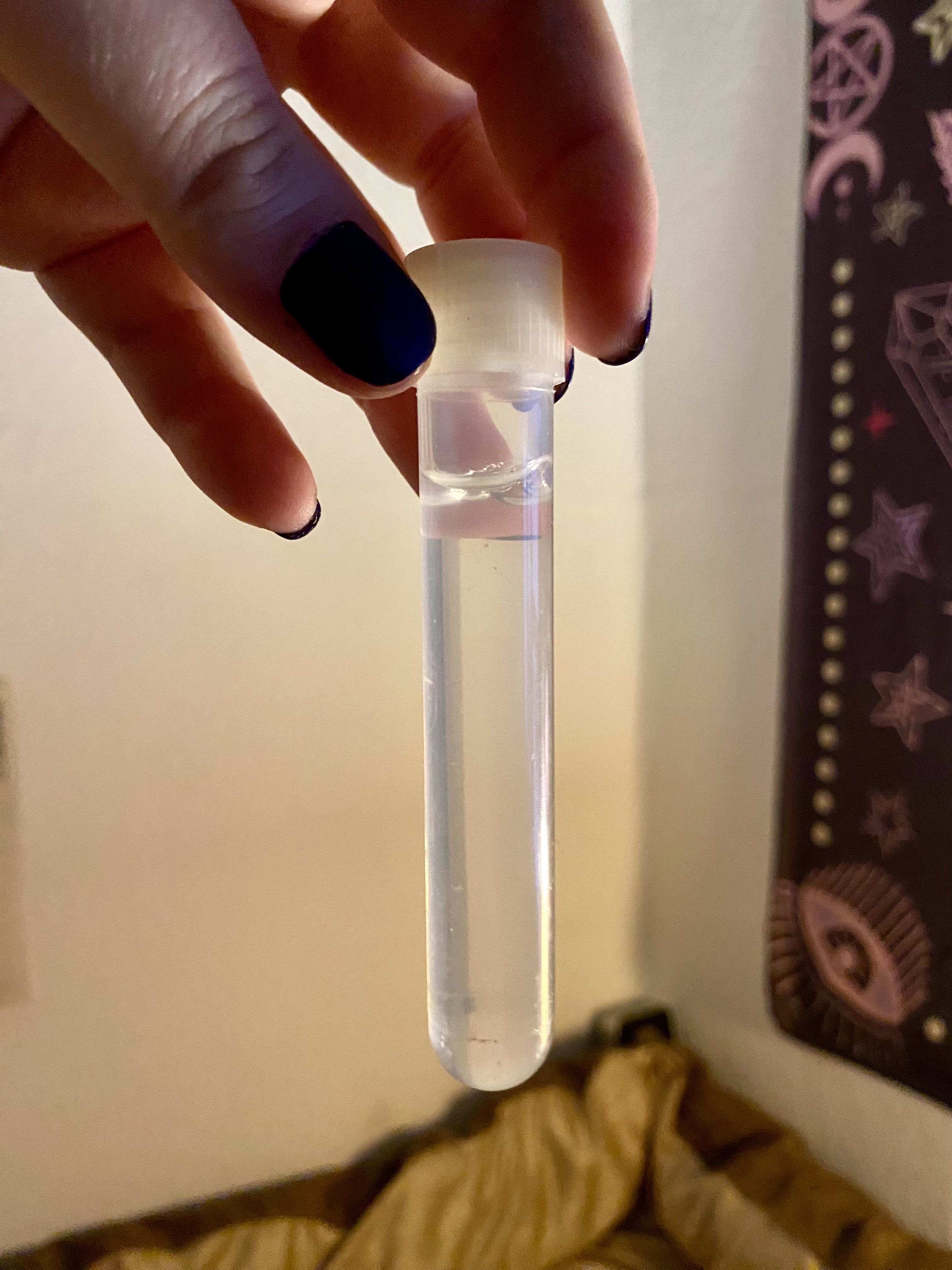 A hand with painted nails holds a small test tube filled with clear liquid. The background is partially visible, showing a patterned wall hanging