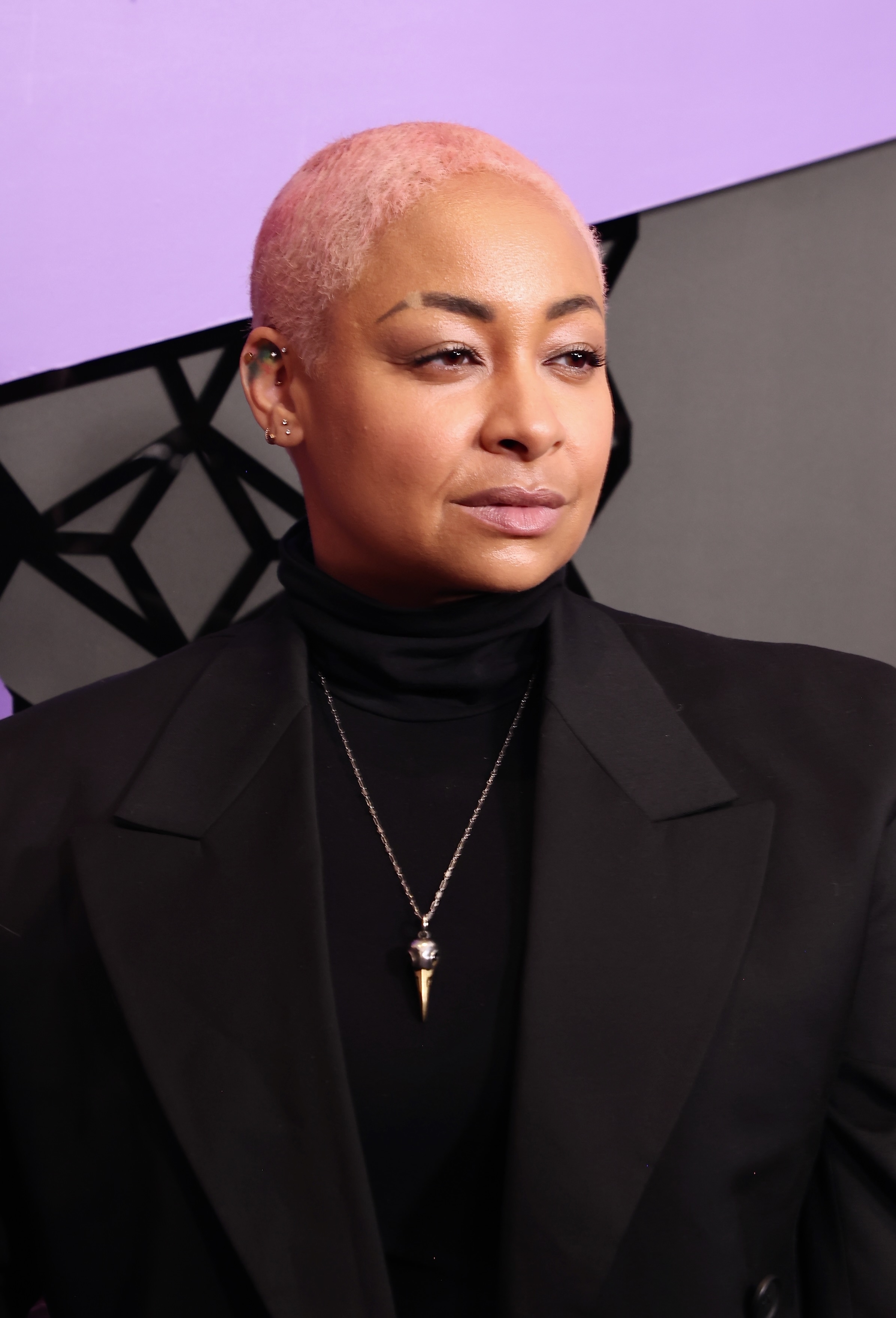 Raven-Symoné with short hair wearing a black turtleneck and statement necklace, posing in front of an abstract background
