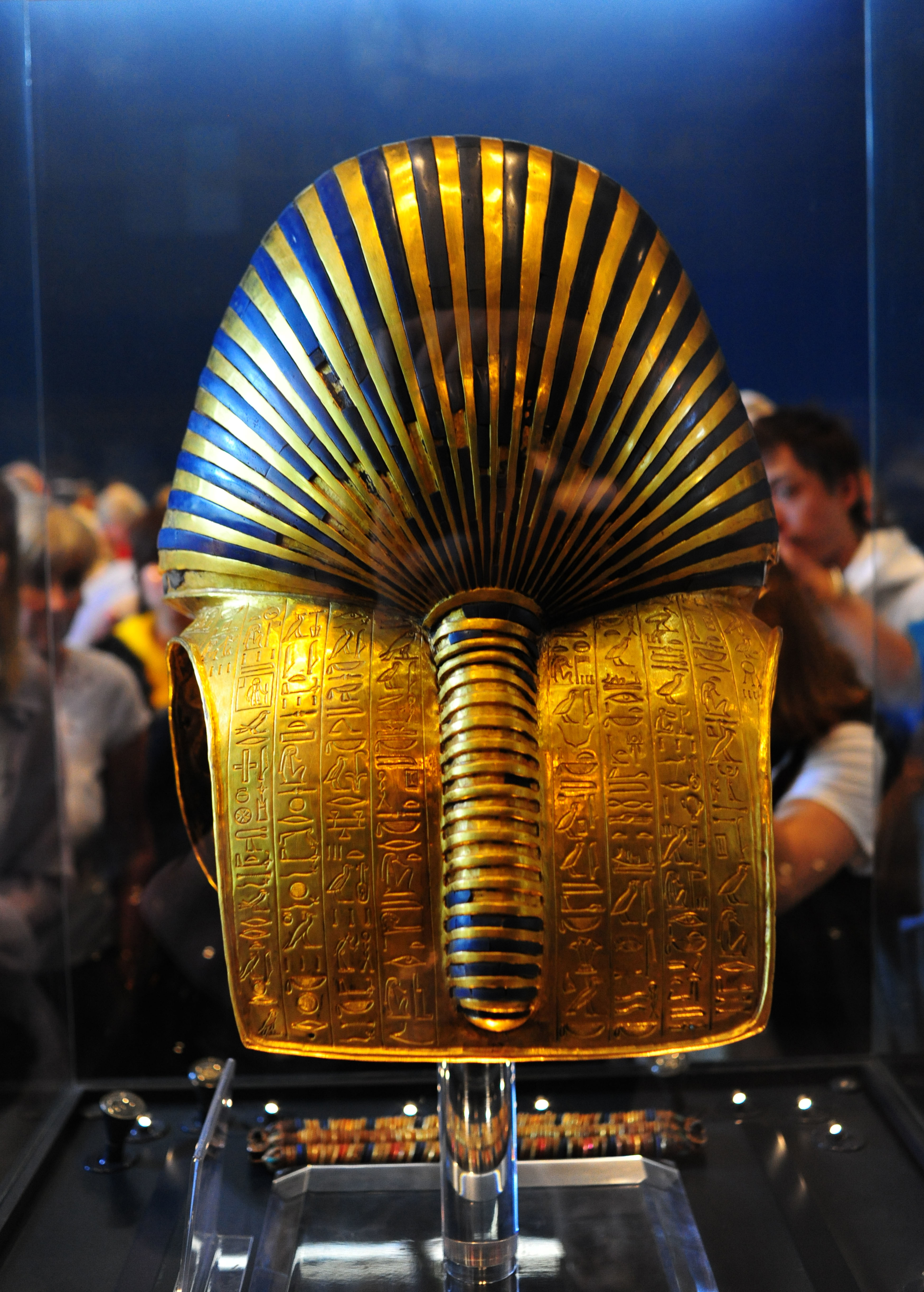 Golden ancient Egyptian pharaoh mask with intricate hieroglyphs on display, viewed from behind with people observing in the background