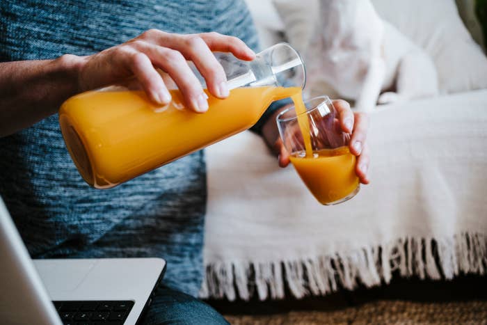 Person pouring a glass of orange juice from a carafe while holding a laptop. They are sitting on a couch with a fringe throw blanket in the background