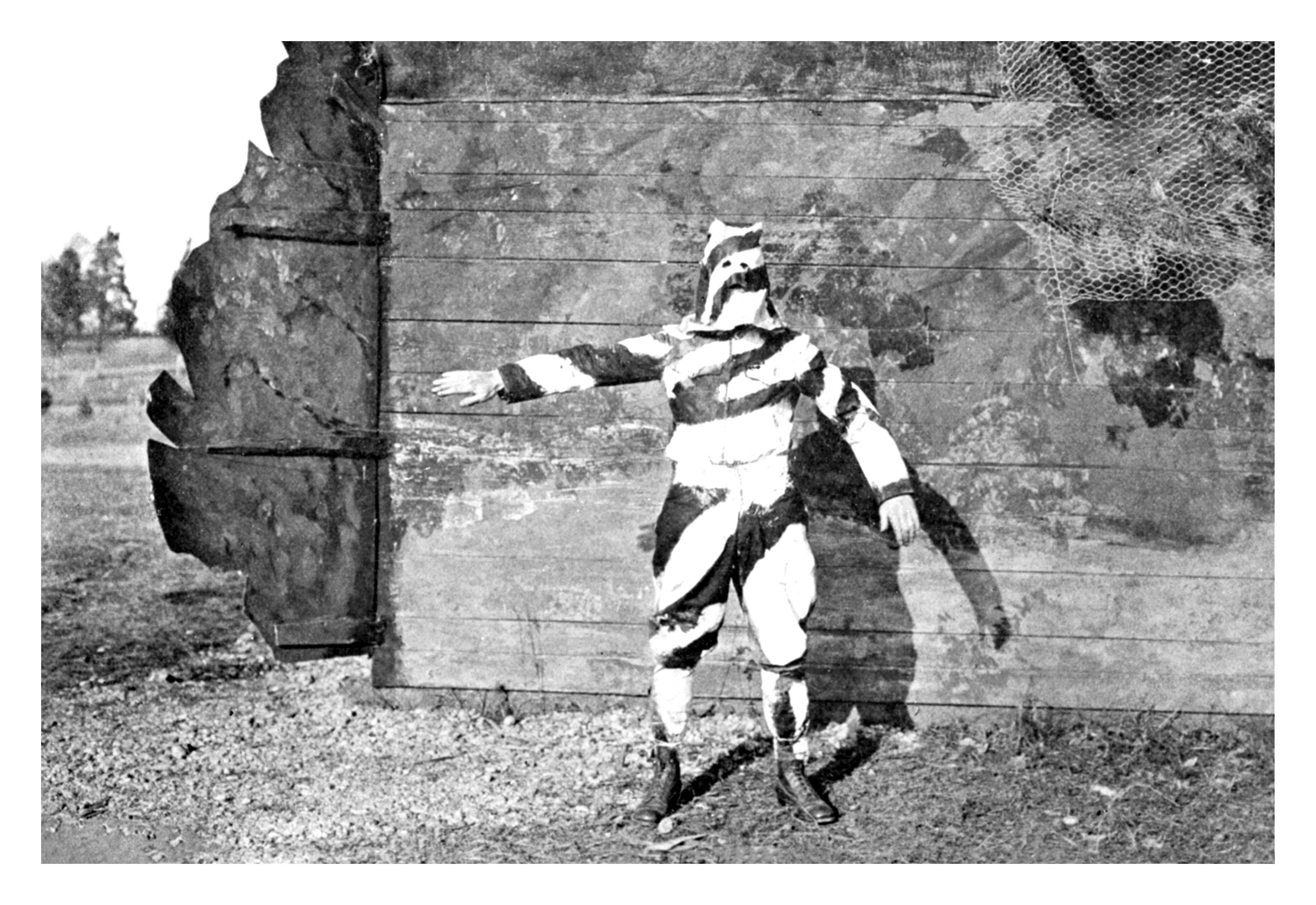 A historical black-and-white photo of a person in a full-body suit with stripes, standing against a backdrop, likely as camouflage. Names unknown