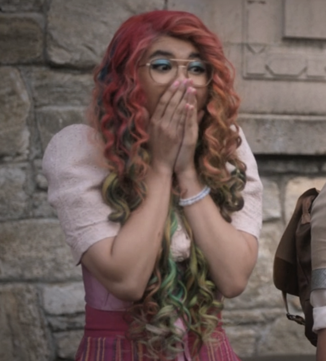 Sam as Zellie, looks surprised with hands covering her mouth. She has wavy hair and wears a puffy-sleeved dress on a stone building backdrop