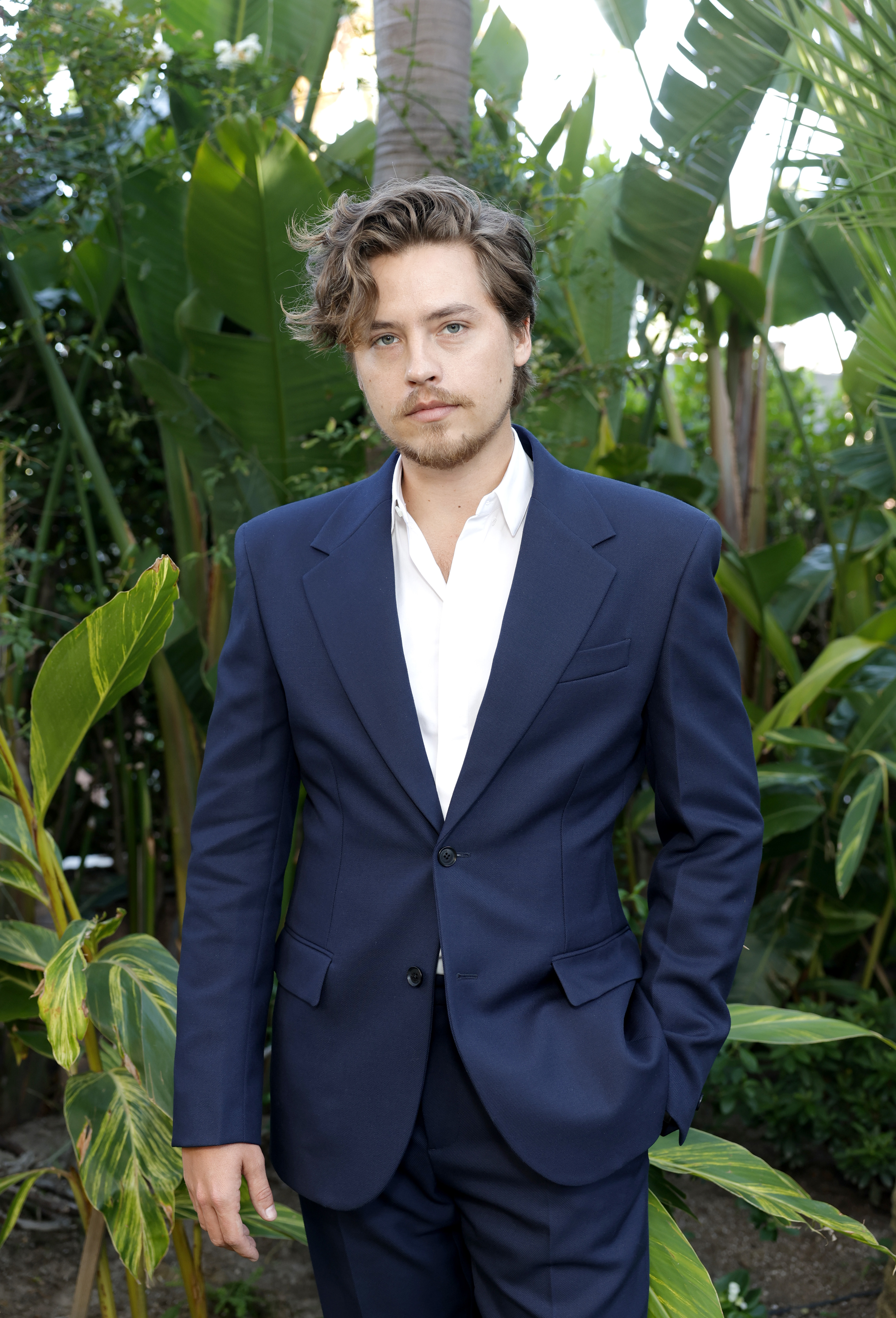 Cole Sprouse stands outdoors amongst lush greenery, wearing a tailored suit over an open-collared shirt