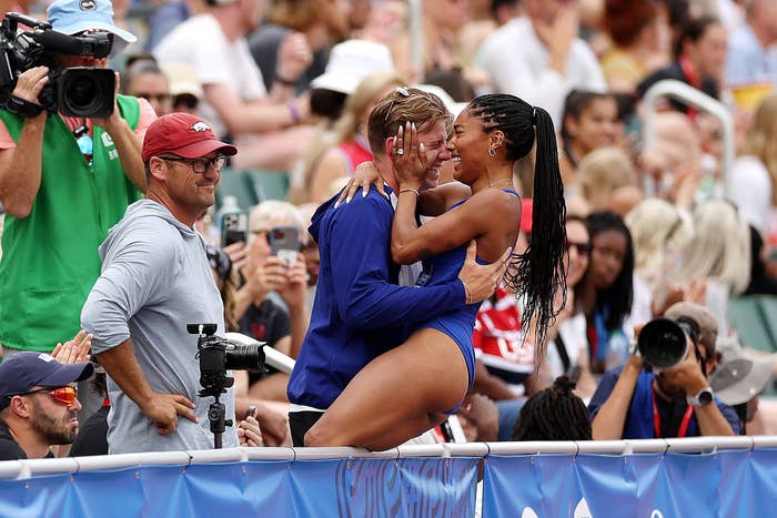 Two athletes celebrate joyfully at a track event, hugging over a barrier, surrounded by a crowd and media photographers