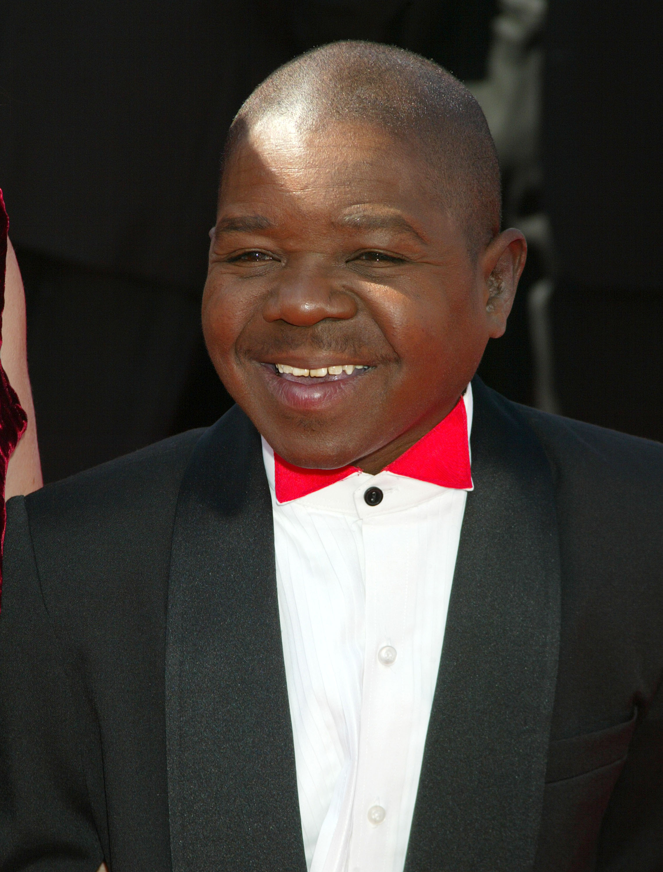 Gary Coleman is smiling at a red carpet event, wearing a black suit with a white shirt and a bright red bow tie