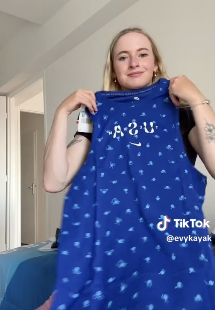 Evy Leibfarth holding up a blue USA-themed tank top with small patterns on it in a bedroom. TikTok watermark and username @evykayak visible