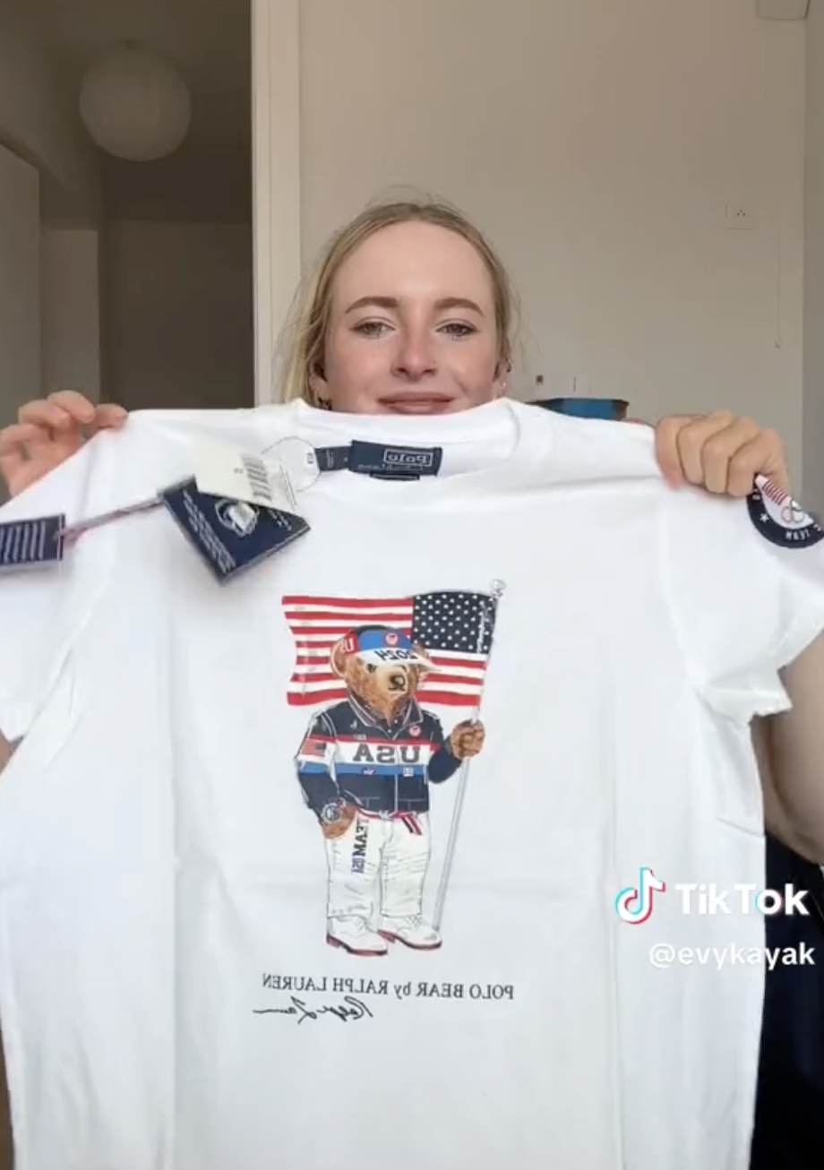 A woman holds up a white Polo Bear by Ralph Lauren T-shirt with a graphic of a bear holding an American flag and wearing a USA jersey. TikTok watermark is seen with username @evykayak