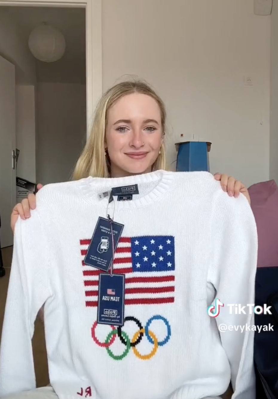 Person smiling and holding up a sweater with an American flag and Olympic rings design. Tags are attached to the sweater. TikTok account @evykayak is visible