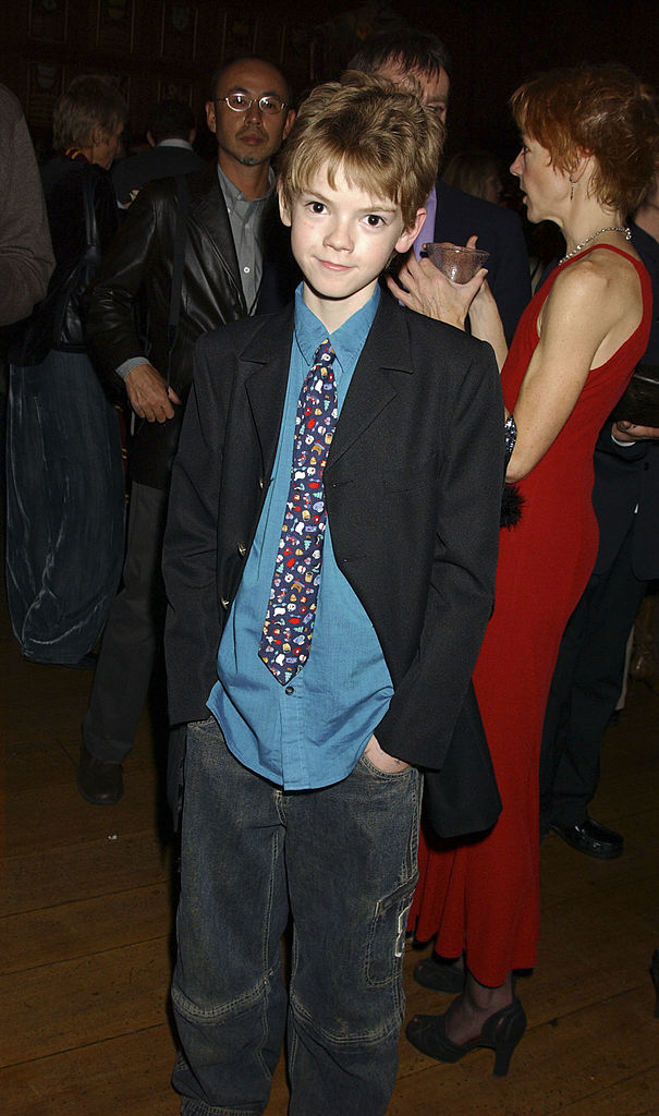Thomas Brodie-Sangster in casual attire at an event, wearing a patterned tie, blue shirt, and black blazer. People converse in the background