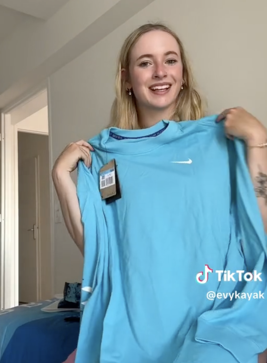 A smiling woman, @evykayak, holds up a new shirt while showcasing it in a TikTok video. She wears casual clothing and is inside a bedroom
