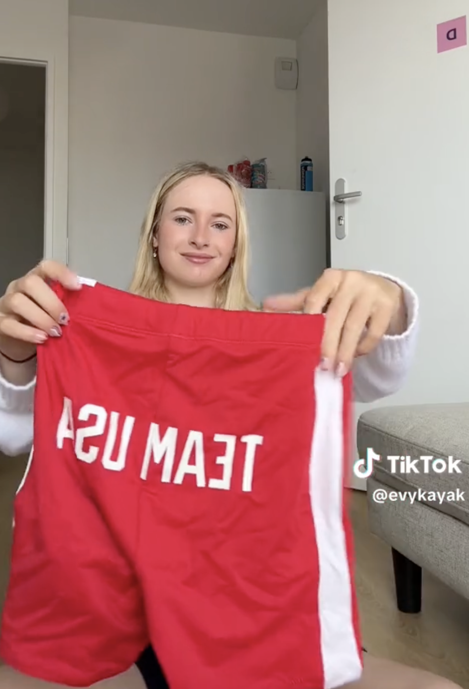 A woman holding red &quot;TEAM USA&quot; shorts, smiling, indoors. TikTok username &quot;@evykayak&quot; displayed