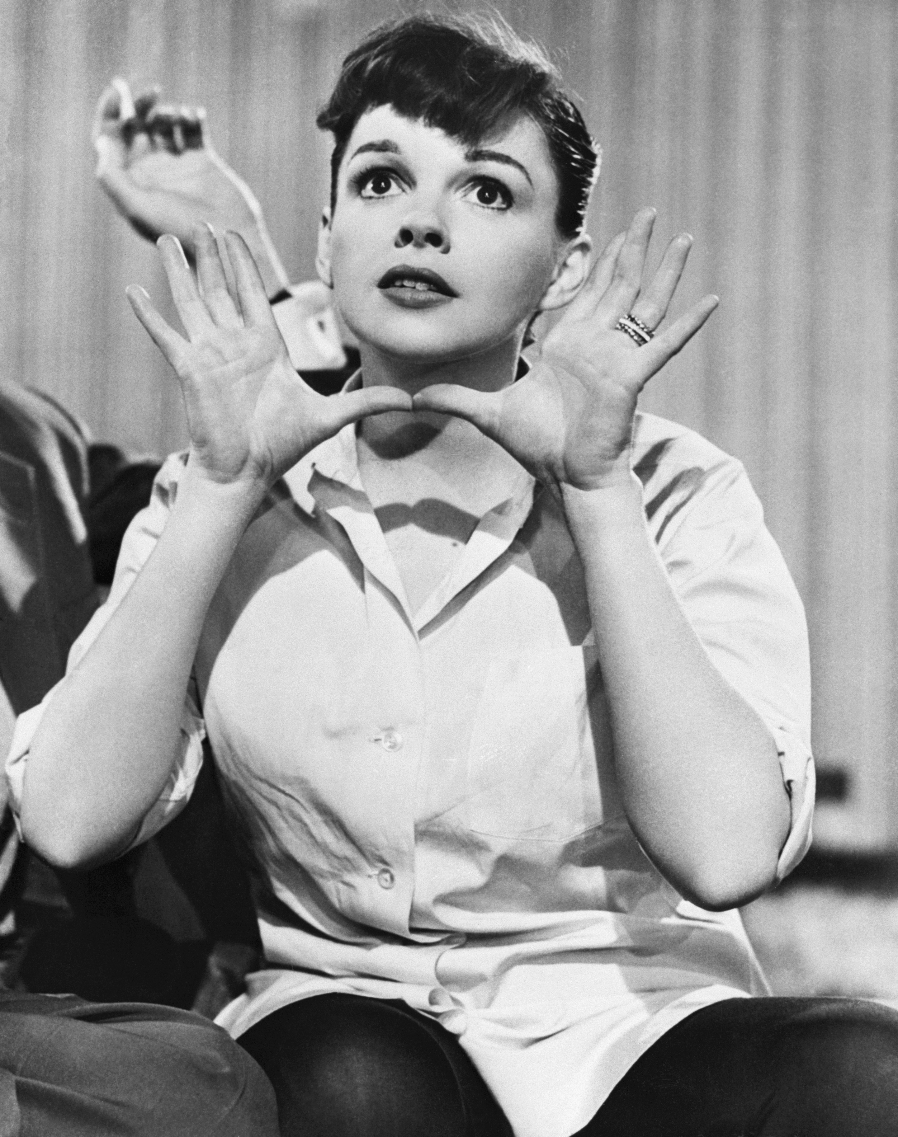 Judy Garland in a white blouse with sleeves rolled up, sitting with hands raised near her face, looking upwards thoughtfully