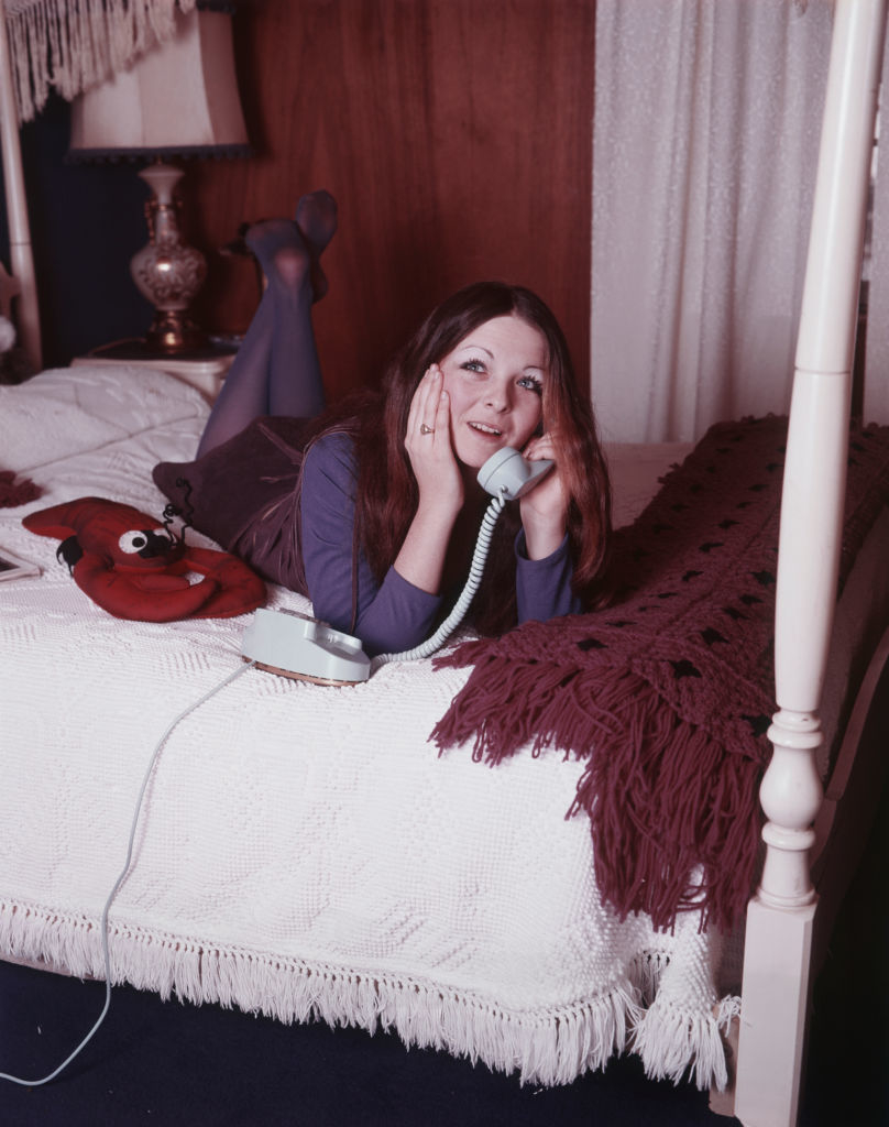 A woman lies on a bed talking on a rotary phone, with a red stuffed toy by her side, in a room with a lamp and crochet bedspread