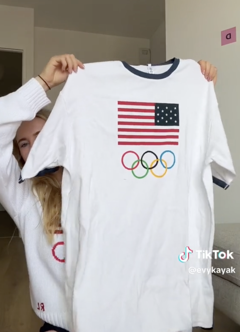 Woman in a white sweater holds up a white T-shirt with the US flag and Olympic rings. TikTok username @evykayak visible