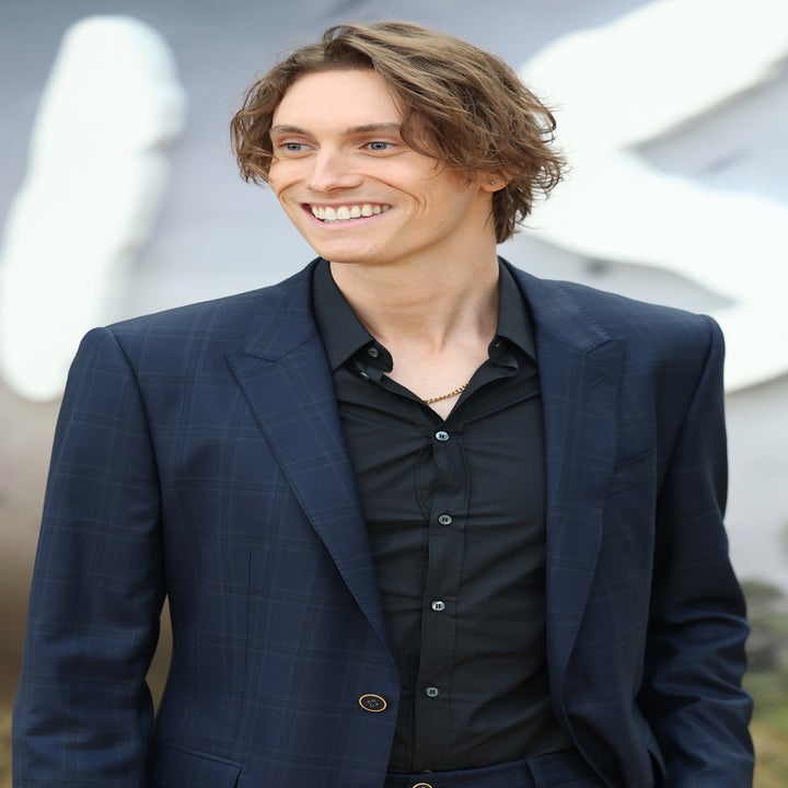Person in a dark suit and black shirt smiling while standing outdoors