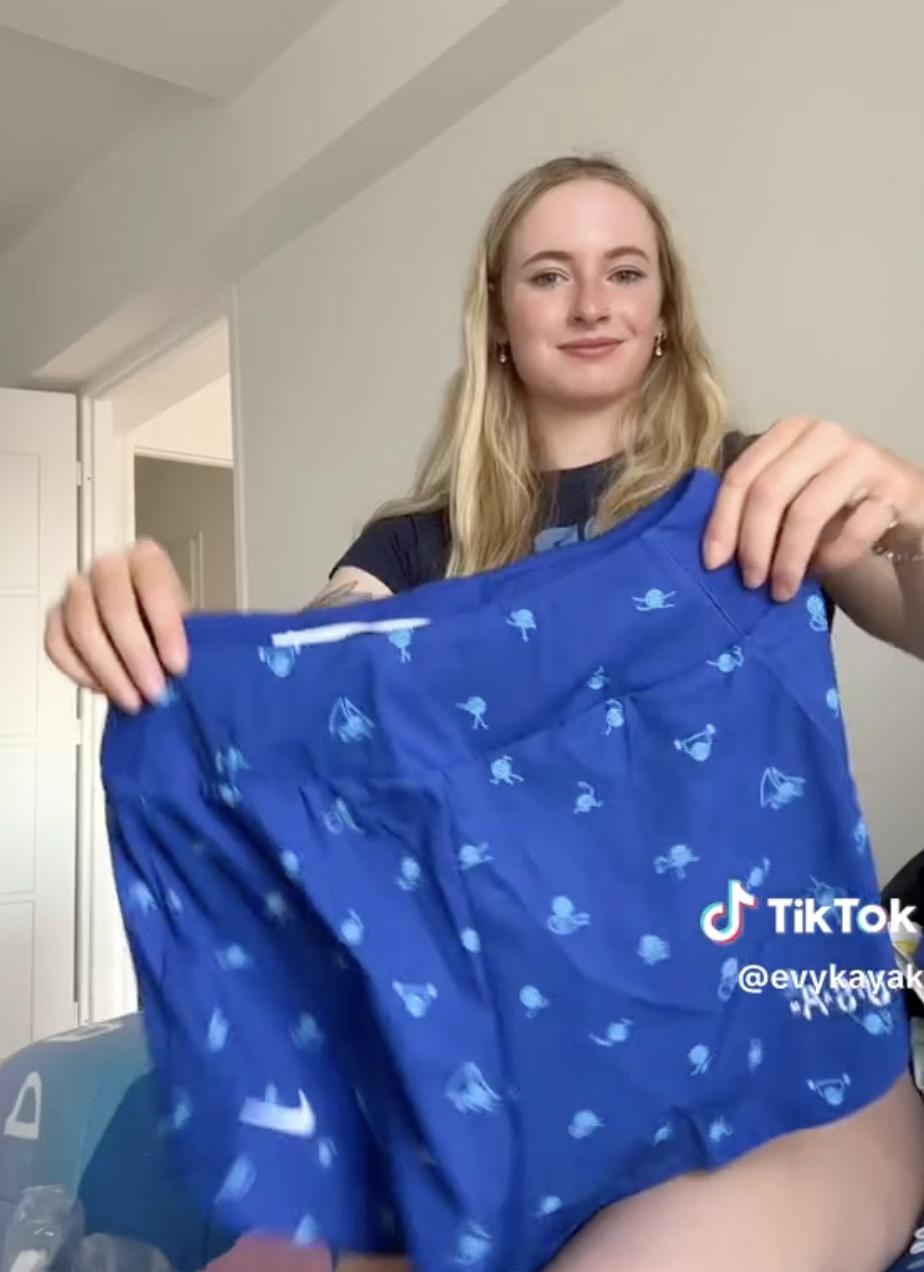 Evy Leibfarth holds up a pair of blue shorts with patterns while smiling at the camera. TikTok username: @evykayak