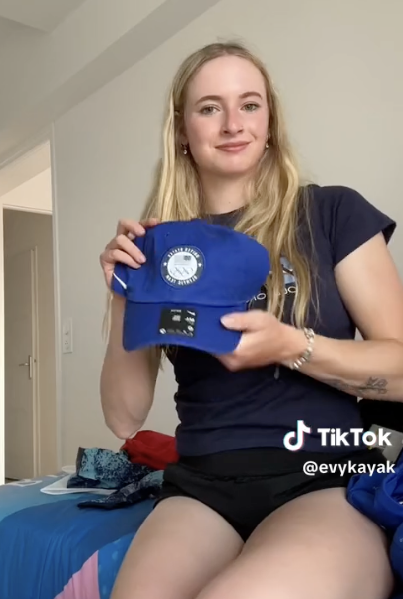 Evy Leibfarth holding a blue cap while sitting on a bed in a casual setting, sporting a casual outfit