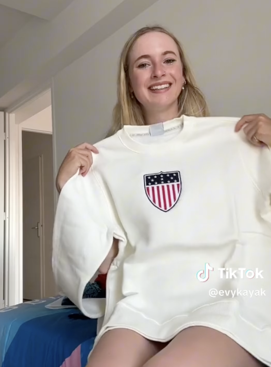 Evykayak smiling and holding up a white sweater with an American flag emblem, in a casual indoor setting. TikTok username @evykayak visible