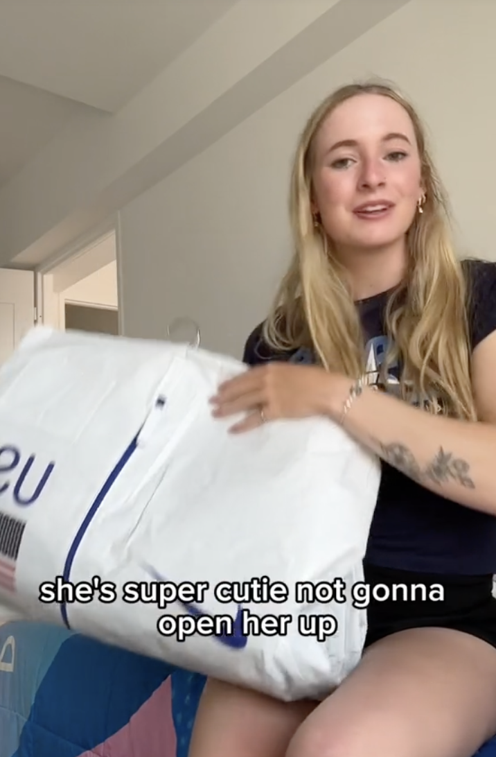 Woman sitting on a bed holding a large white package, with text: &quot;she's super cutie not gonna open her up.&quot;