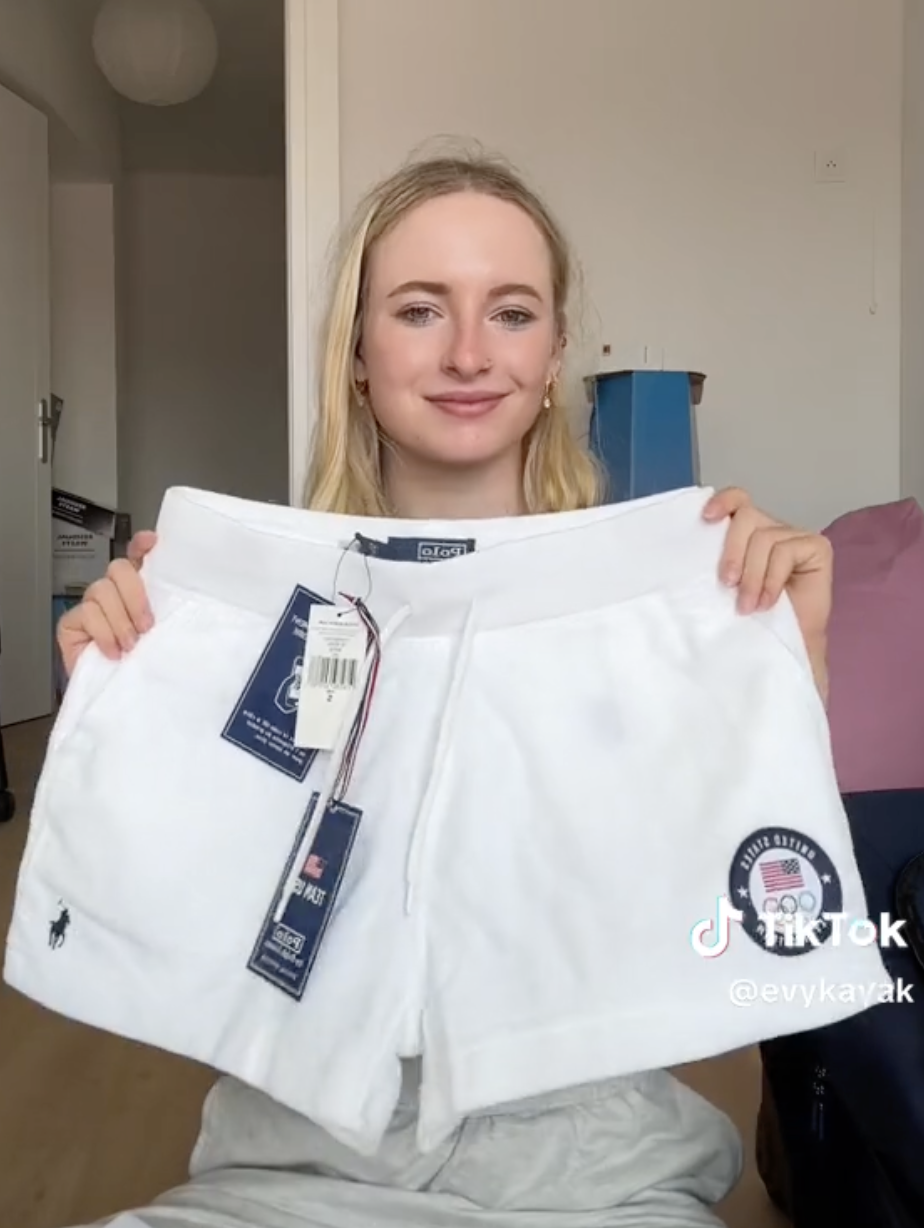Evy Kayaki holds up white shorts with tags. She smiles, with a room visible in the background