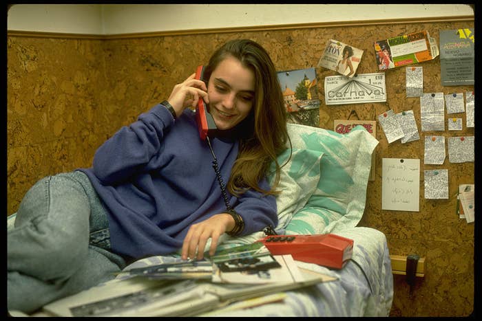 Young woman relaxing on a bed while talking on a corded phone, surrounded by photos and notes on the wall. She looks at a photo album in her lap