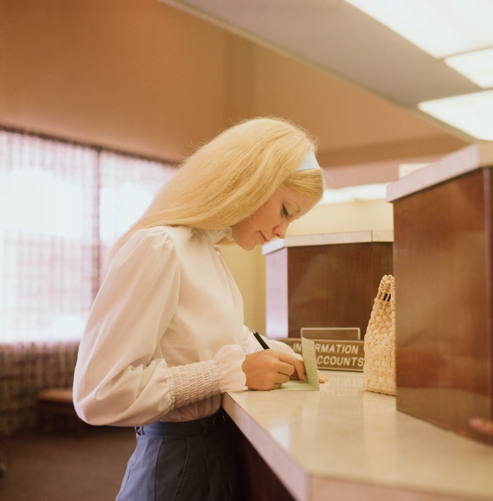 A woman with long, straight hair wearing a white blouse and high-waisted pants is writing at a bank counter in a 1960s-style setting