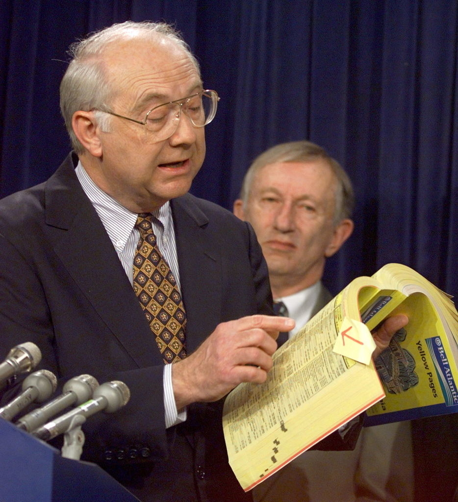 I don't know who these people are. A man in a suit and tie holds an open phone book, pointing to a page, while speaking at a podium with microphones. Another man stands behind him