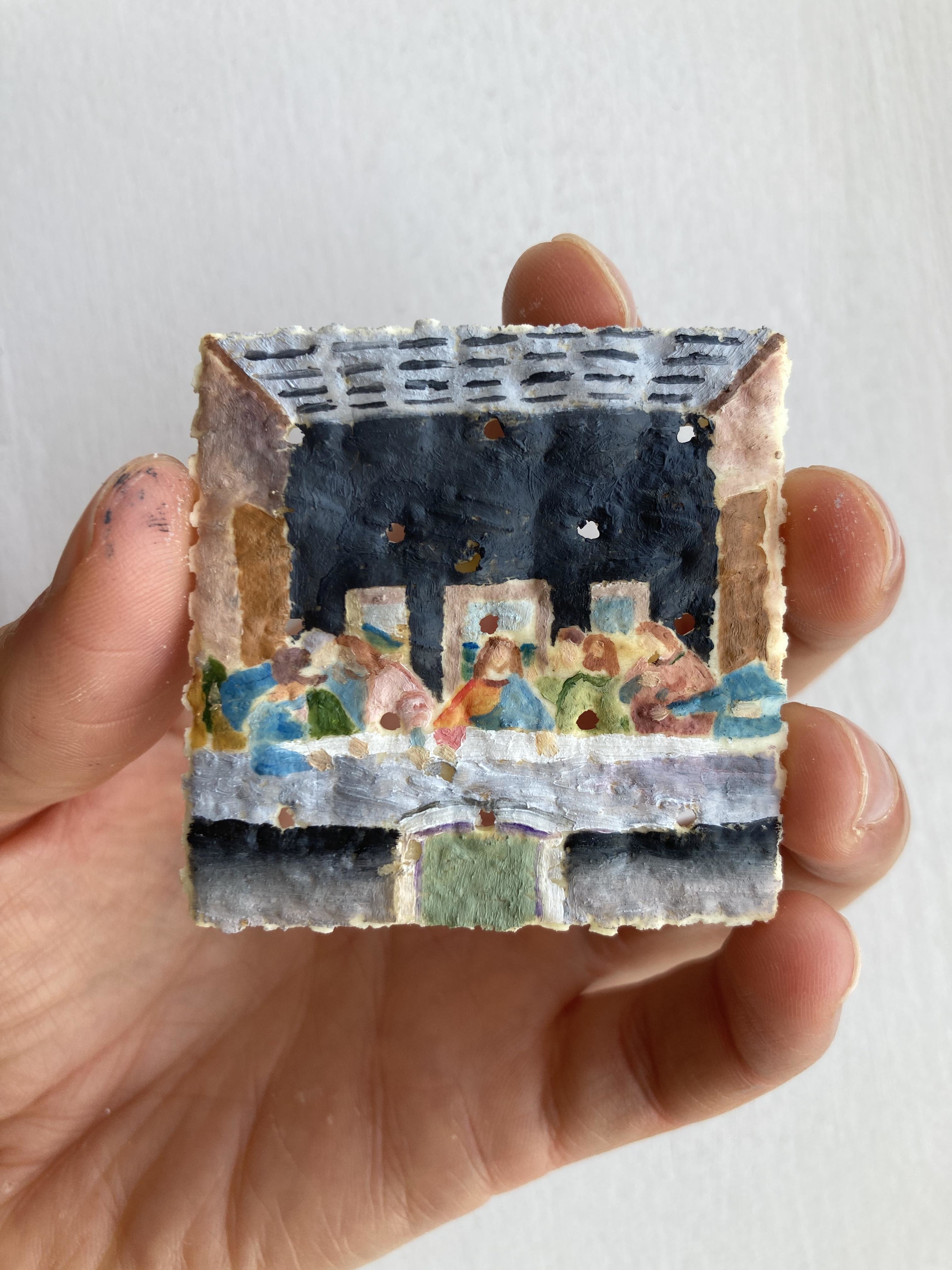 A hand holding a small, square piece of food art depicting the Last Supper scene. The detailed artwork shows figures similar to those in the famous painting
