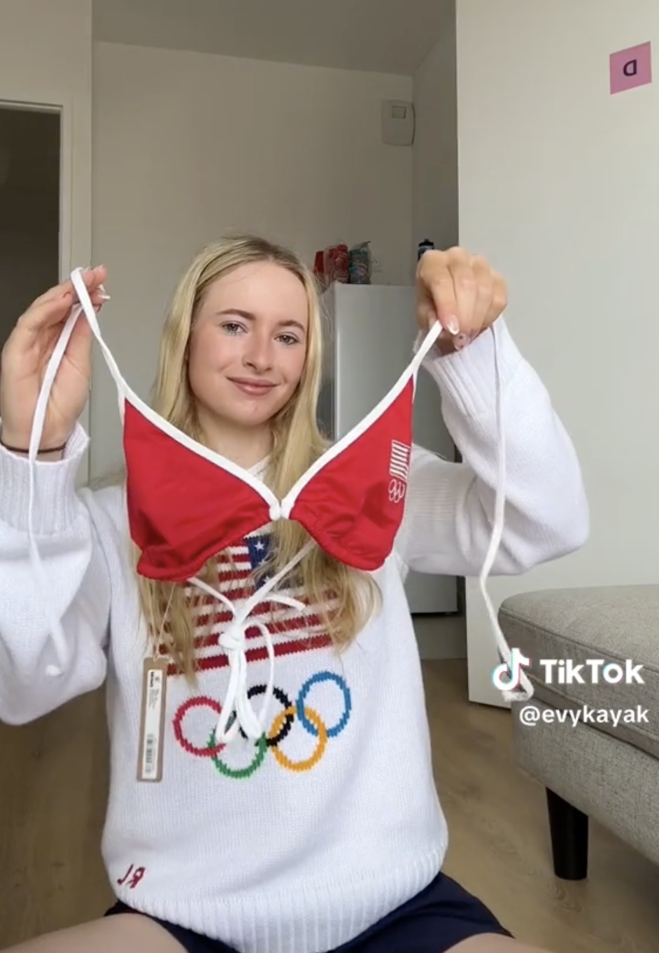 Evy Leibfarth holding a red bikini top and smiling in a room. She is wearing a sweater with the Olympic rings. TikTok handle @evykayak is visible on the image