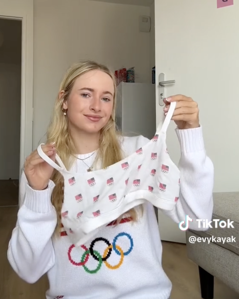 Young woman wearing an Olympic-themed sweater holds up a baby tank top with a printed design. TikTok username @evykayak is visible