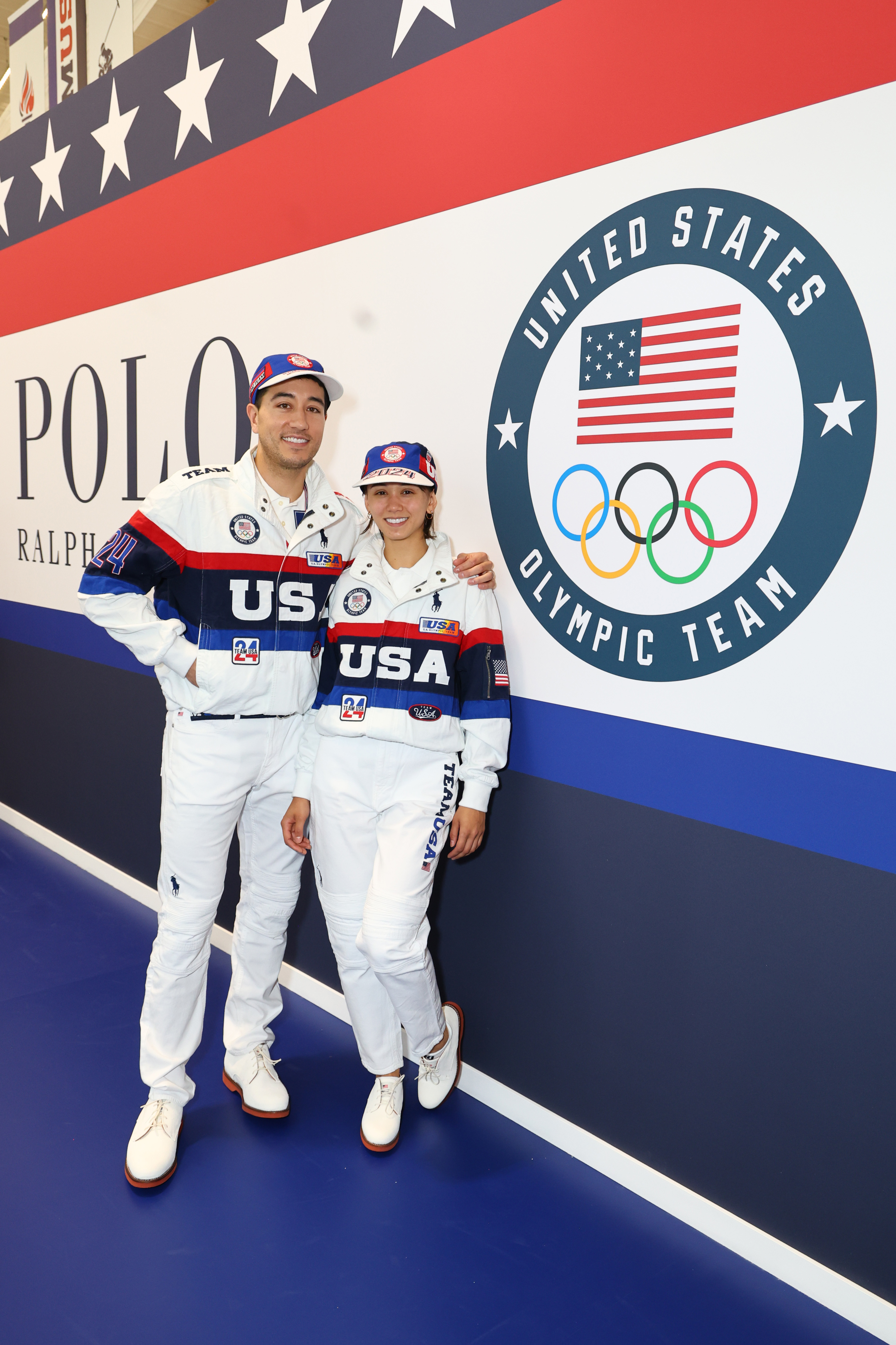 Nathan Chen and Chloe Kim in white USA Olympic tracksuits and hats, standing together, posing near a United States Olympic Team banner