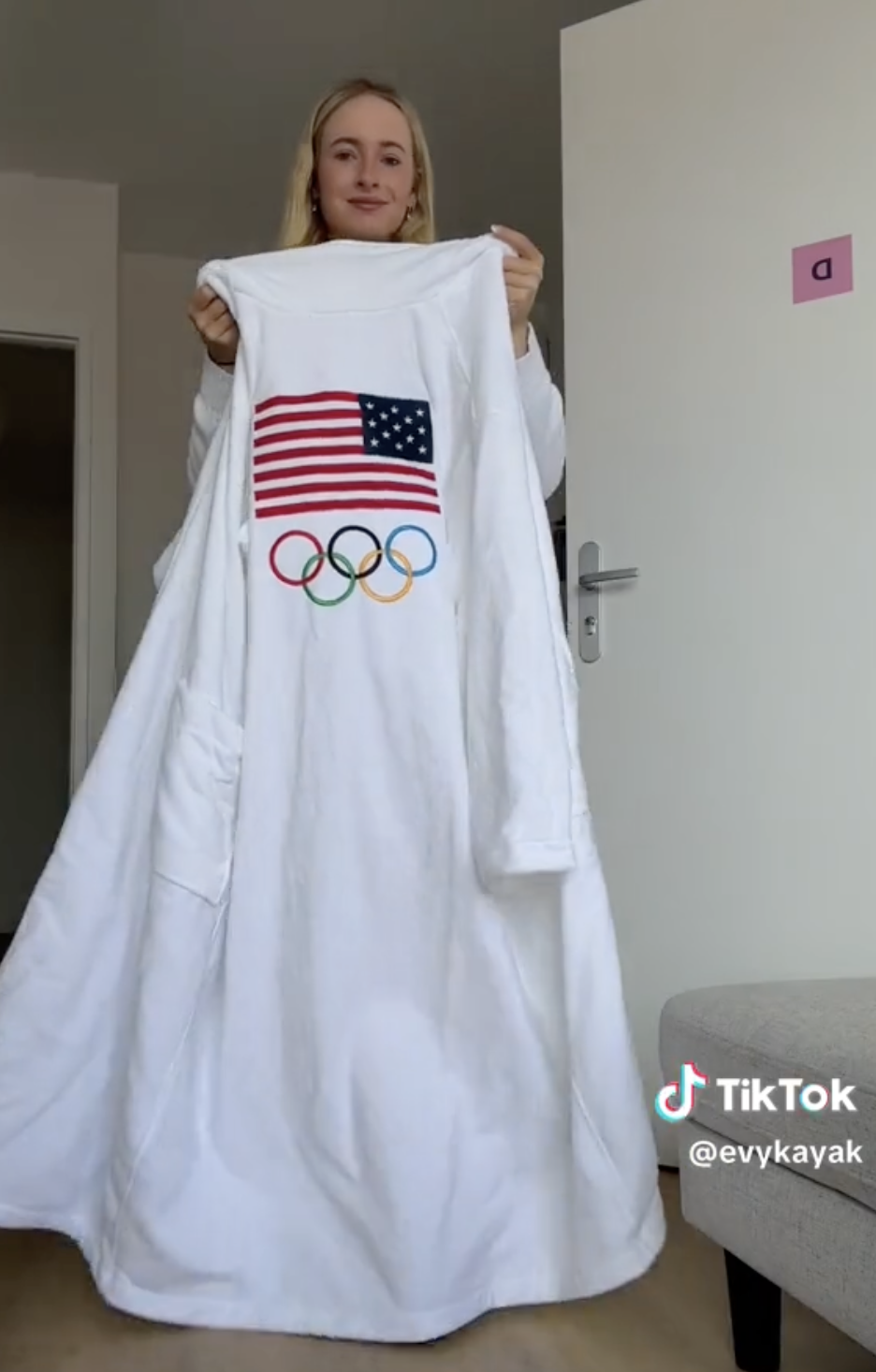 Evy Leibfarth holds up a long white robe with the US flag and Olympic rings on it inside a room. TikTok username @evykayak is visible