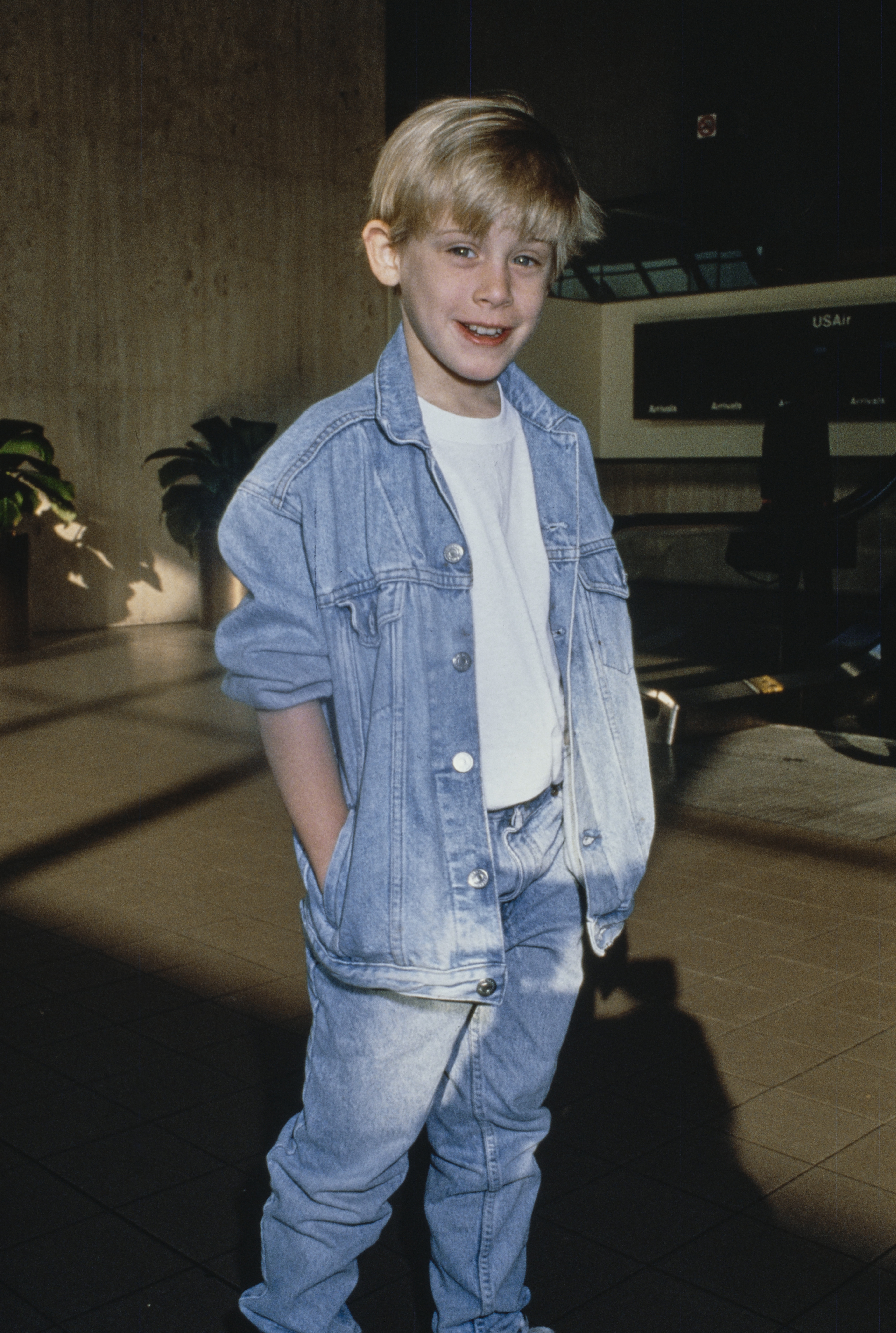 A young boy wearing a denim jacket and jeans with hands in his pockets stands indoors, smiling